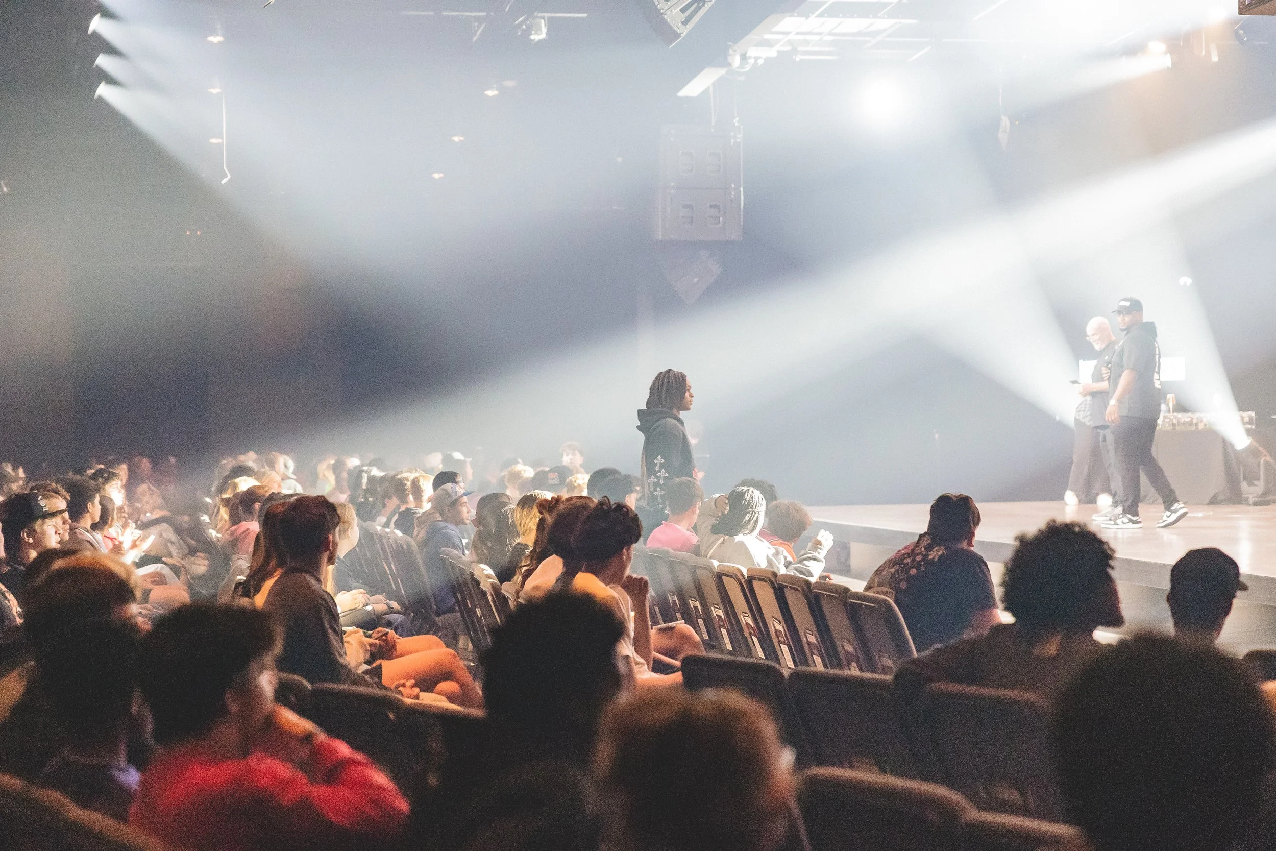 Audience seated in a dark theater watching a stage performance with bright white lights and two people standing at the front.