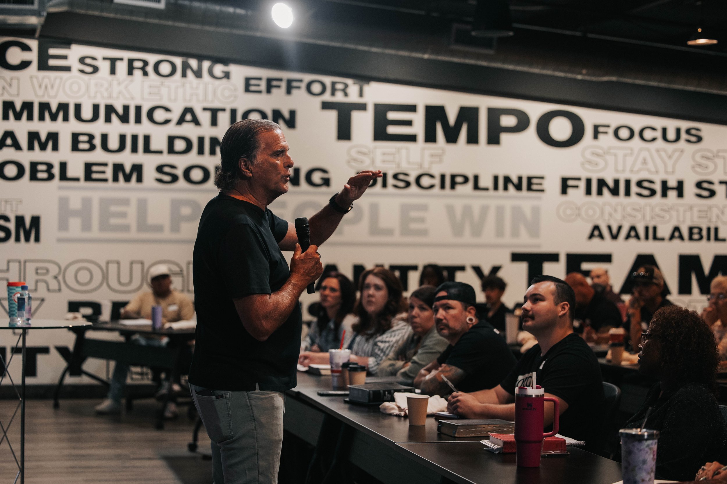 A man speaking into a microphone in a classroom or seminar room filled with people, with a wall covered in motivational words in the background