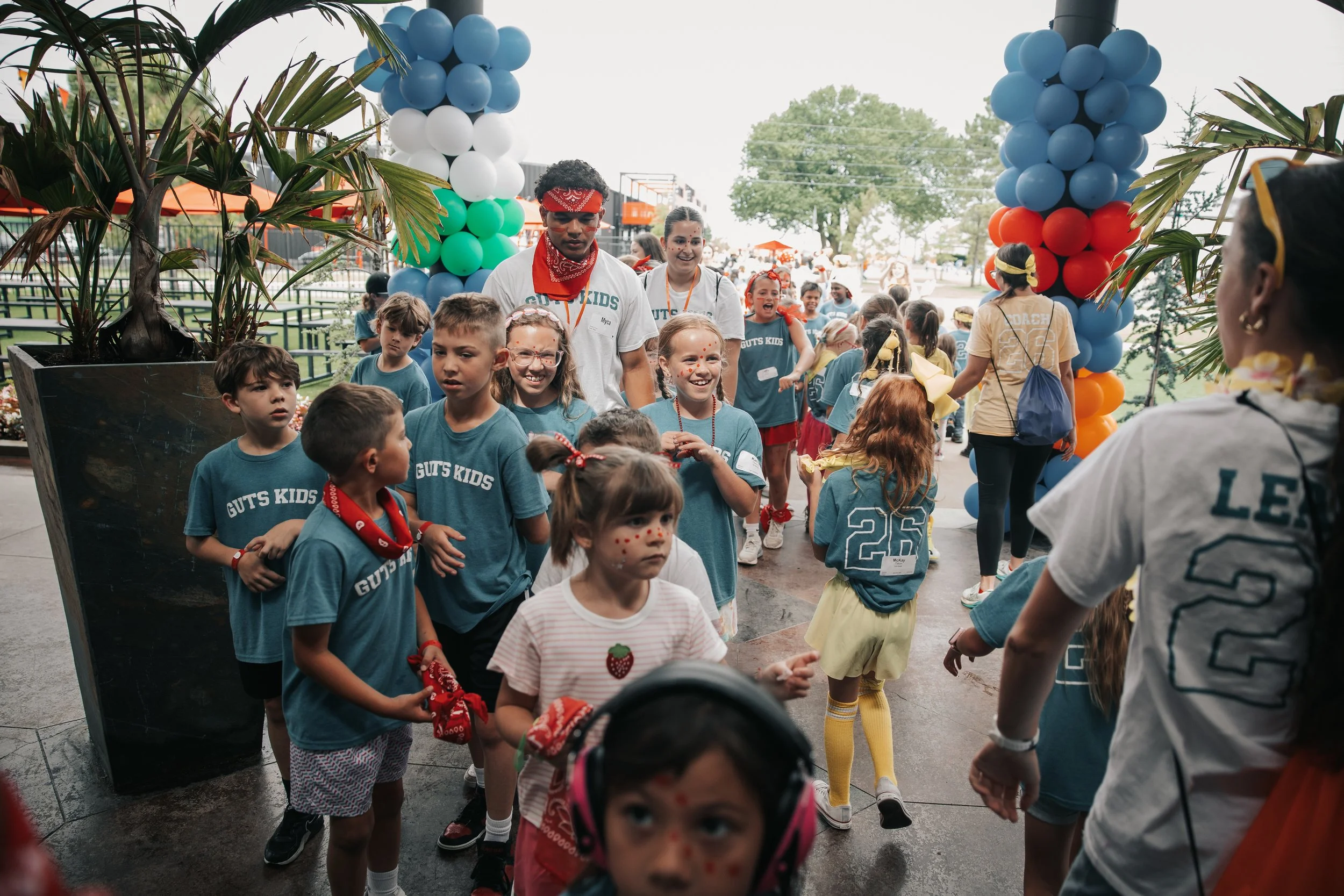 Group of children and adults gathered outdoors at a celebration, decorated with colorful balloon pillars, wearing casual and themed clothing, some with face paint, under a partly cloudy sky.