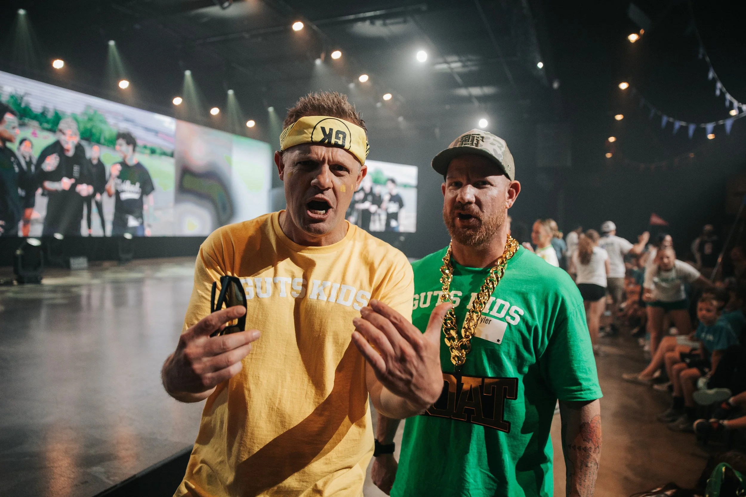 Two men wearing colorful T-shirts with 'GUTS KIDS' printed on them, standing in an indoor venue with a crowd and large screens in the background. The man on the left has a yellow headband and facial paint, with his mouth open, gesturing with his hand