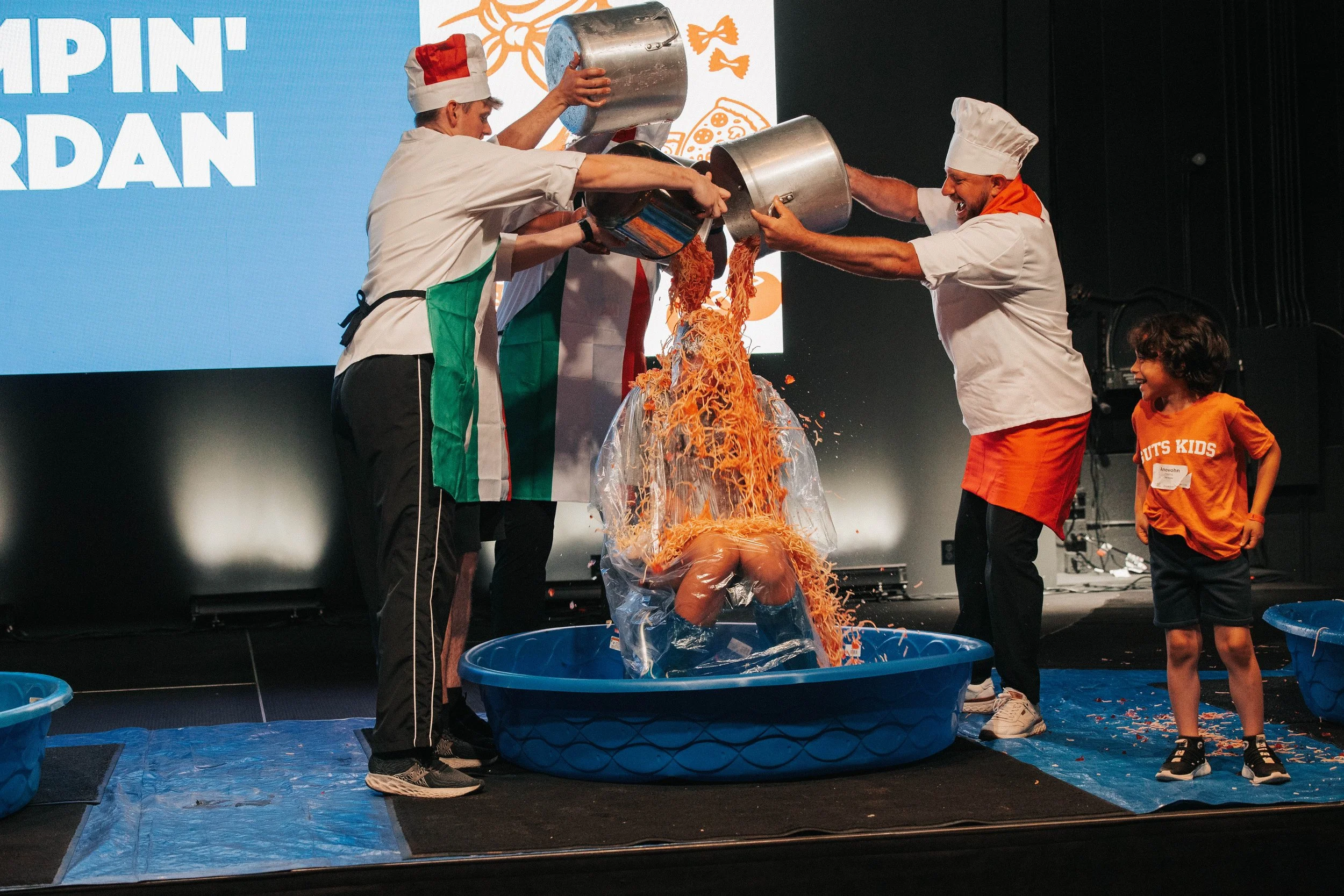 People dressed as chefs pouring noodles into a plastic bag over a person kneeling in a blue tub, with a young girl watching and smiling.