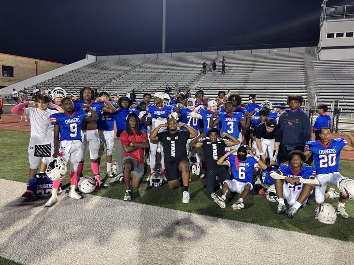 A youth football team in blue jerseys and white pants posing for a group photo on a football field at night, with some players standing and others kneeling, in front of empty bleachers.