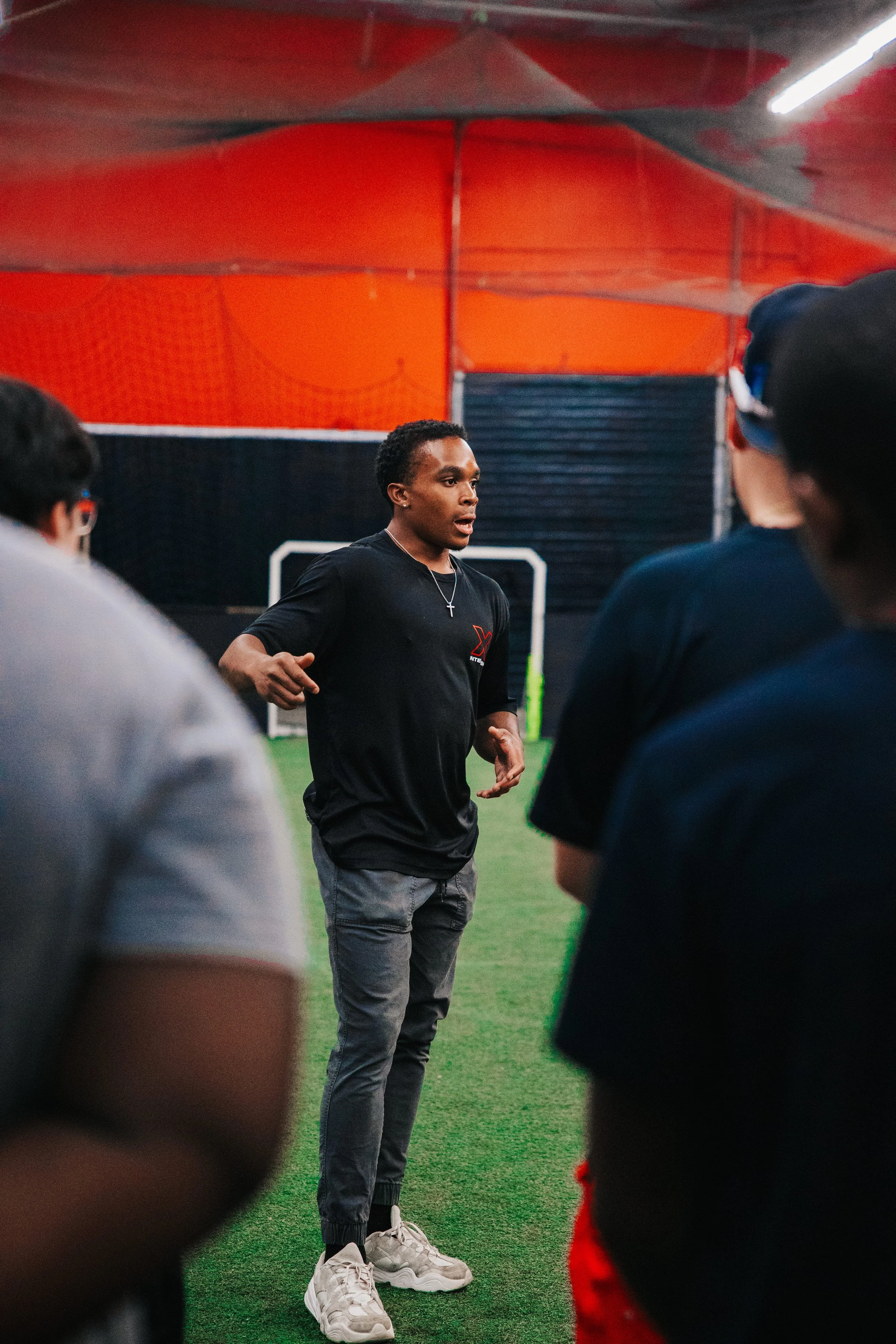A young man giving a speech or instructions to a group of people in an indoor sports facility with green artificial turf and orange and black walls.