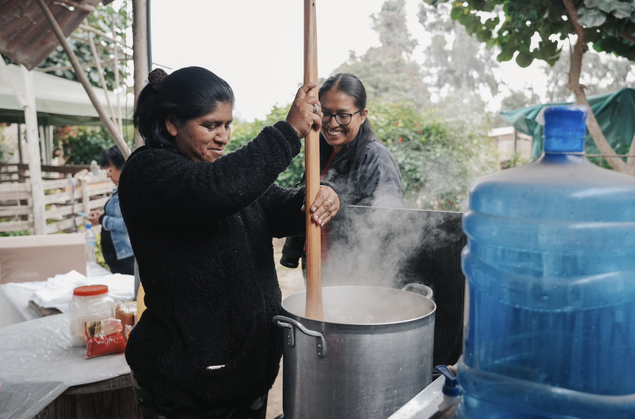 Woman stirring a large pot of boiling food outdoors, smiling, with another woman watching, in a garden setting.