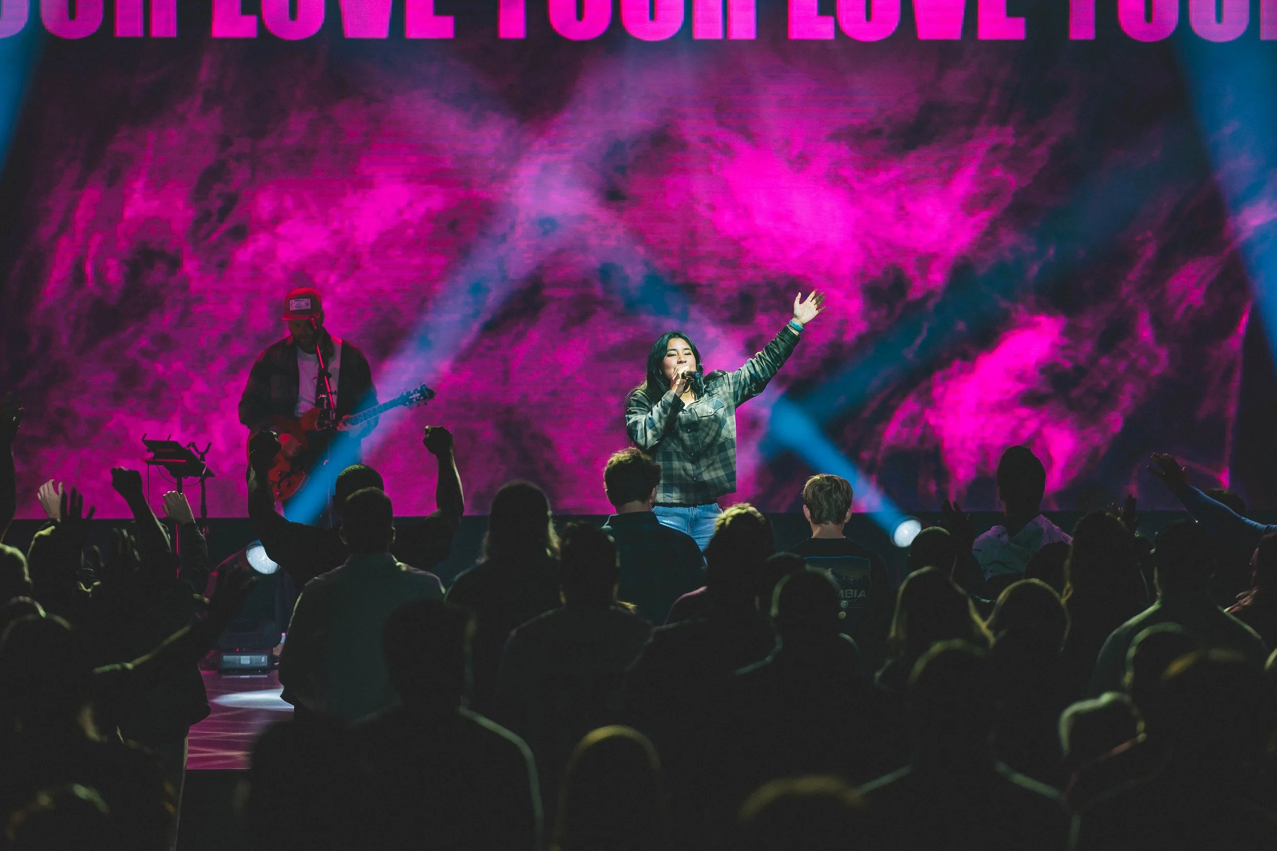 A woman is singing on stage with a microphone, surrounded by an audience. She is raising one arm, and a musician playing guitar is nearby. The background features a vivid pink and purple cosmic pattern and the words "LOVE YOU" in large capital letters at the top.