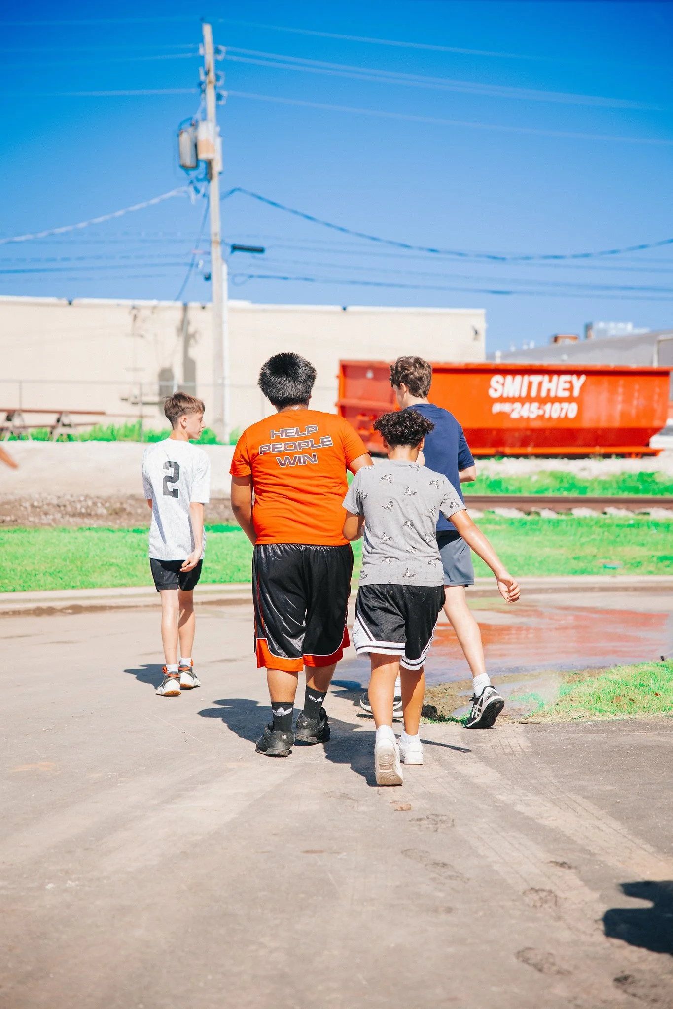 Group of children walking on a dirt path outdoors in a sunny area, with a red freight train behind them and a blue sky overhead.