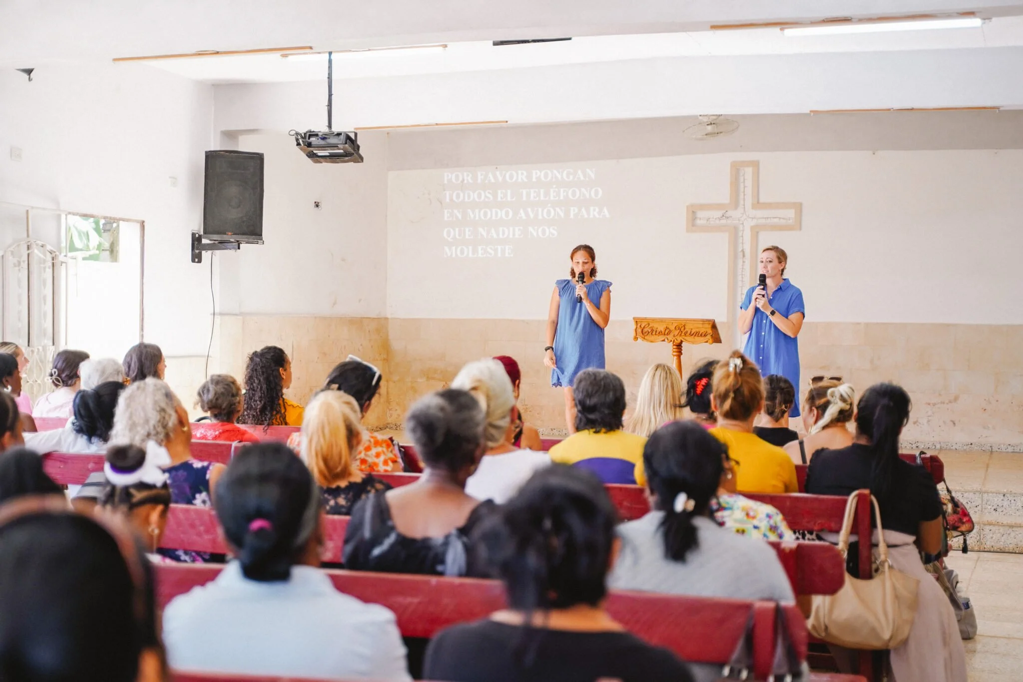A church gathering with two women on stage holding microphones, addressing an audience of diverse women, with a cross on the wall behind them and a projected text in Spanish.