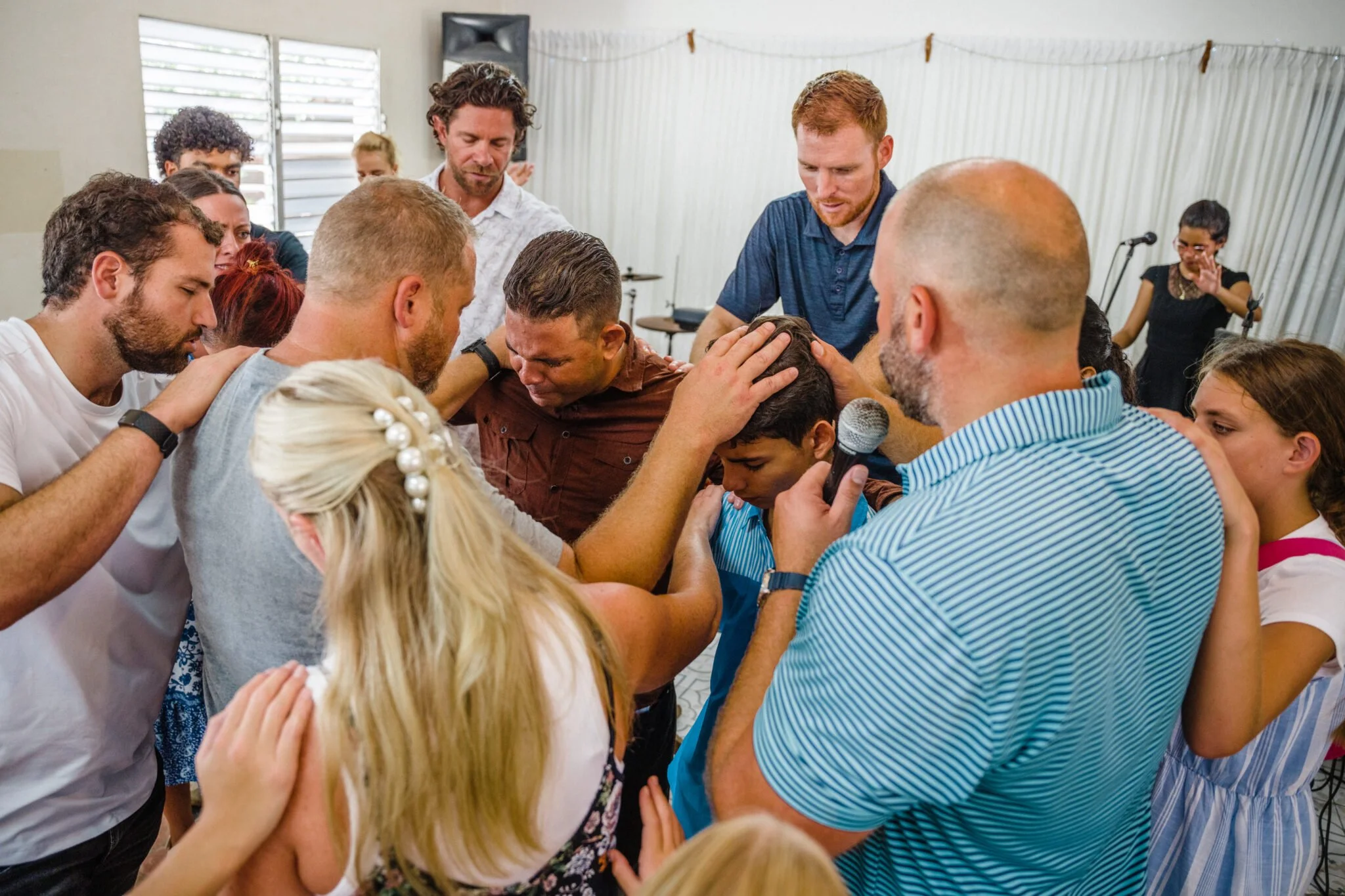 A group of people gathered closely together in prayer, with some having their eyes closed and heads bowed, in an indoor setting with white curtains and windows.
