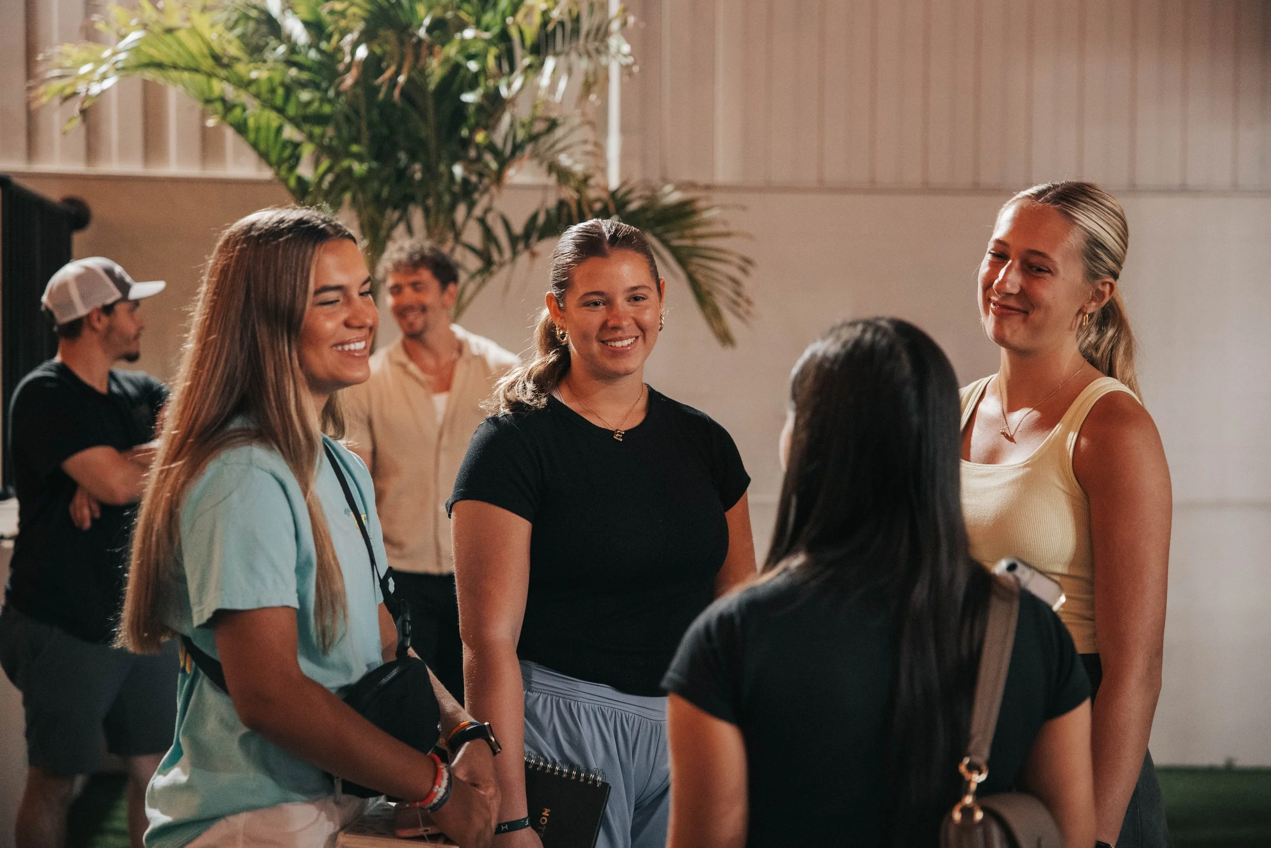 Four young women are talking and smiling together indoors, with a tropical plant in the background and two young men in the background.