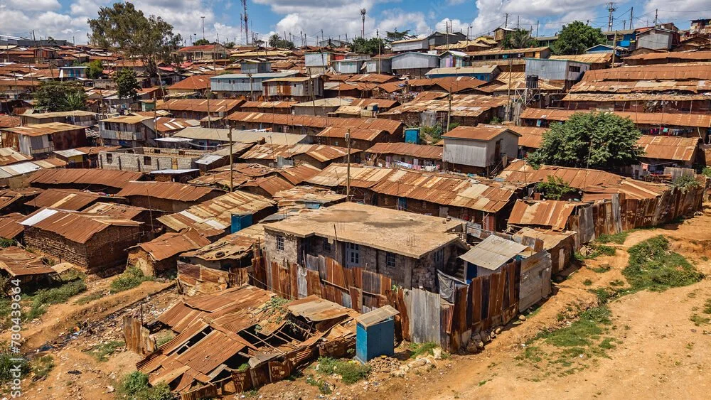 A densely packed hillside neighborhood with makeshift houses made of rusted metal sheets and wood, with dirt paths and sparse greenery.