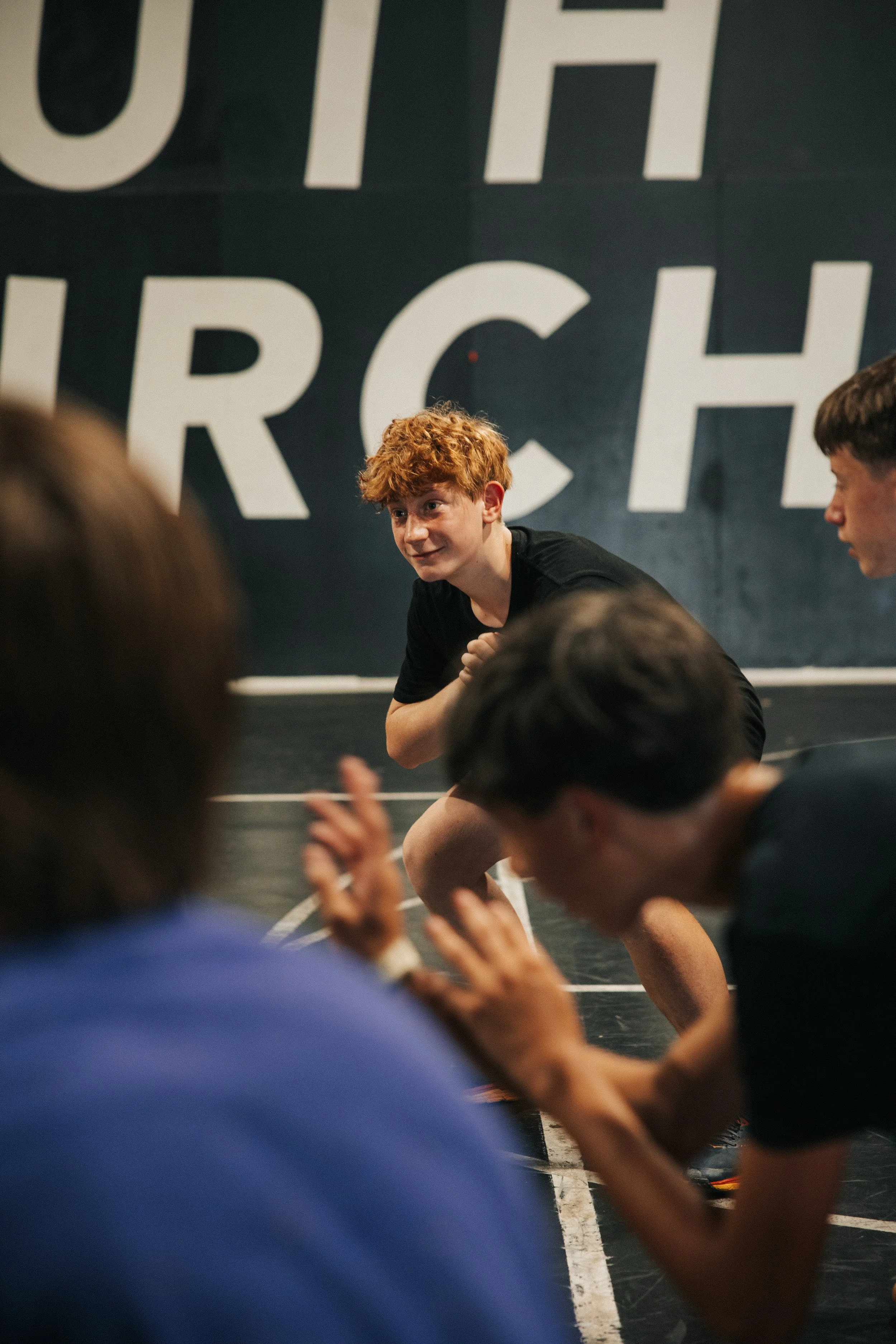 A young man with curly red hair in a black shirt is practicing wrestling moves on a mat, surrounded by other individuals in an indoor gym or training facility.