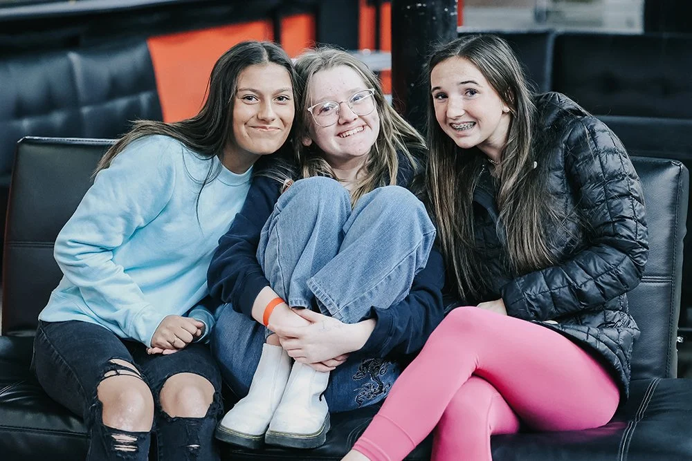 Three teenage girls sitting on a black couch, smiling and enjoying each other's company.