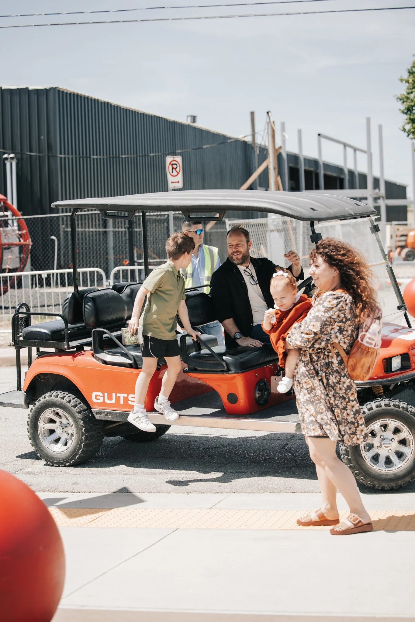 A family stands near a small orange utility vehicle labeled 'GUTS' at an outdoor location. Two children are sitting on the vehicle; one child is talking to a man sitting inside, and the other is being held by a woman with curly hair. The scene is bright and sunny, with a black building, a fence, and a 'No Parking' sign in the background.