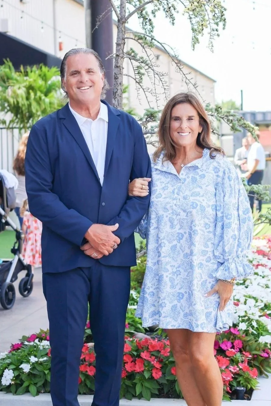 A middle-aged man in a navy blue suit and a woman in a light blue floral dress smiling and standing outside near a small tree and flower garden.