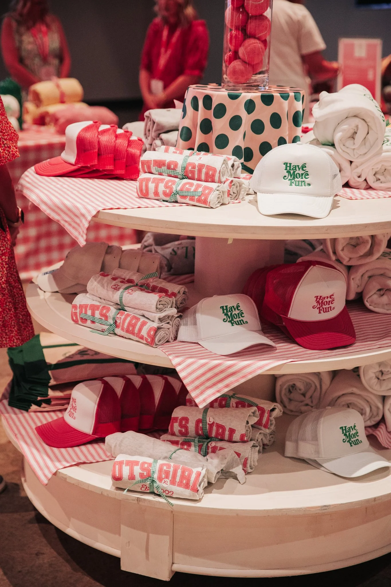 Display of red and white caps, rolled towels, and folded shirts on a round wooden table, with a glass jar of pink candies on top. The items feature phrases like 'Have More Fun' and 'Girls Guts,' and are arranged with red and pink accents in a retail 