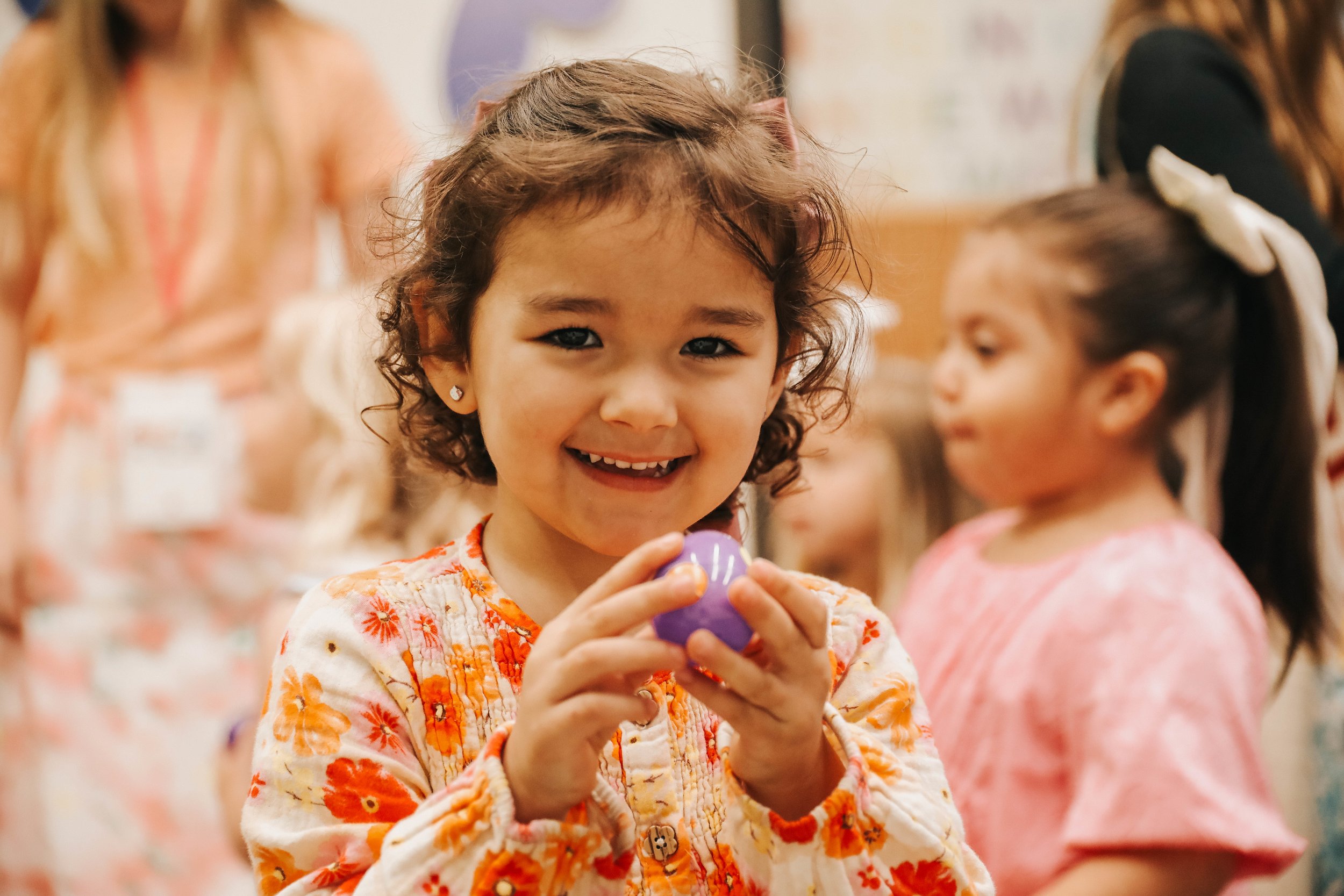 A young girl with curly hair holding a purple Easter egg and smiling at the camera, with other children in the background.