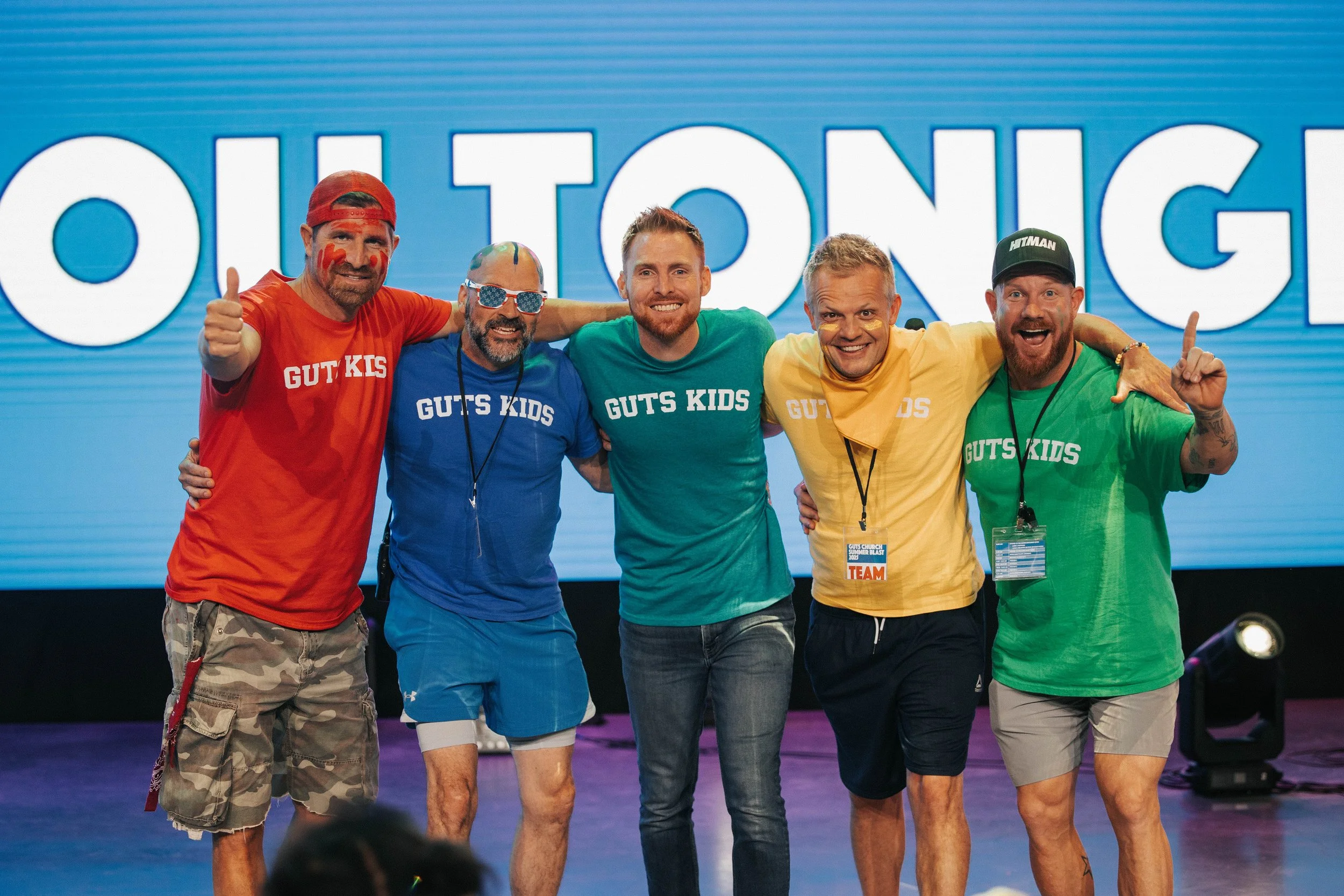 Group of five men standing on stage, wearing colorful T-shirts with 'GUTS KIDS' printed on them, smiling and posing with their arms around each other, with a large screen displaying partially visible text in the background.
