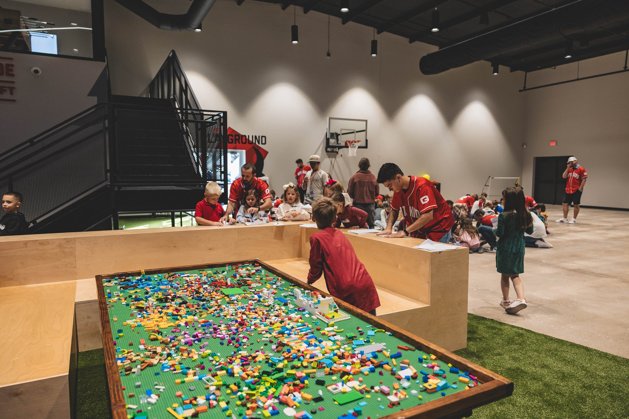 Children playing with building blocks on a large green Lego table in an indoor kids' play area at a sports or entertainment center.