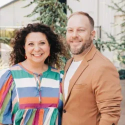 A smiling woman with curly hair and a man with a beard, both standing outdoors in front of greenery, posing for a photo.