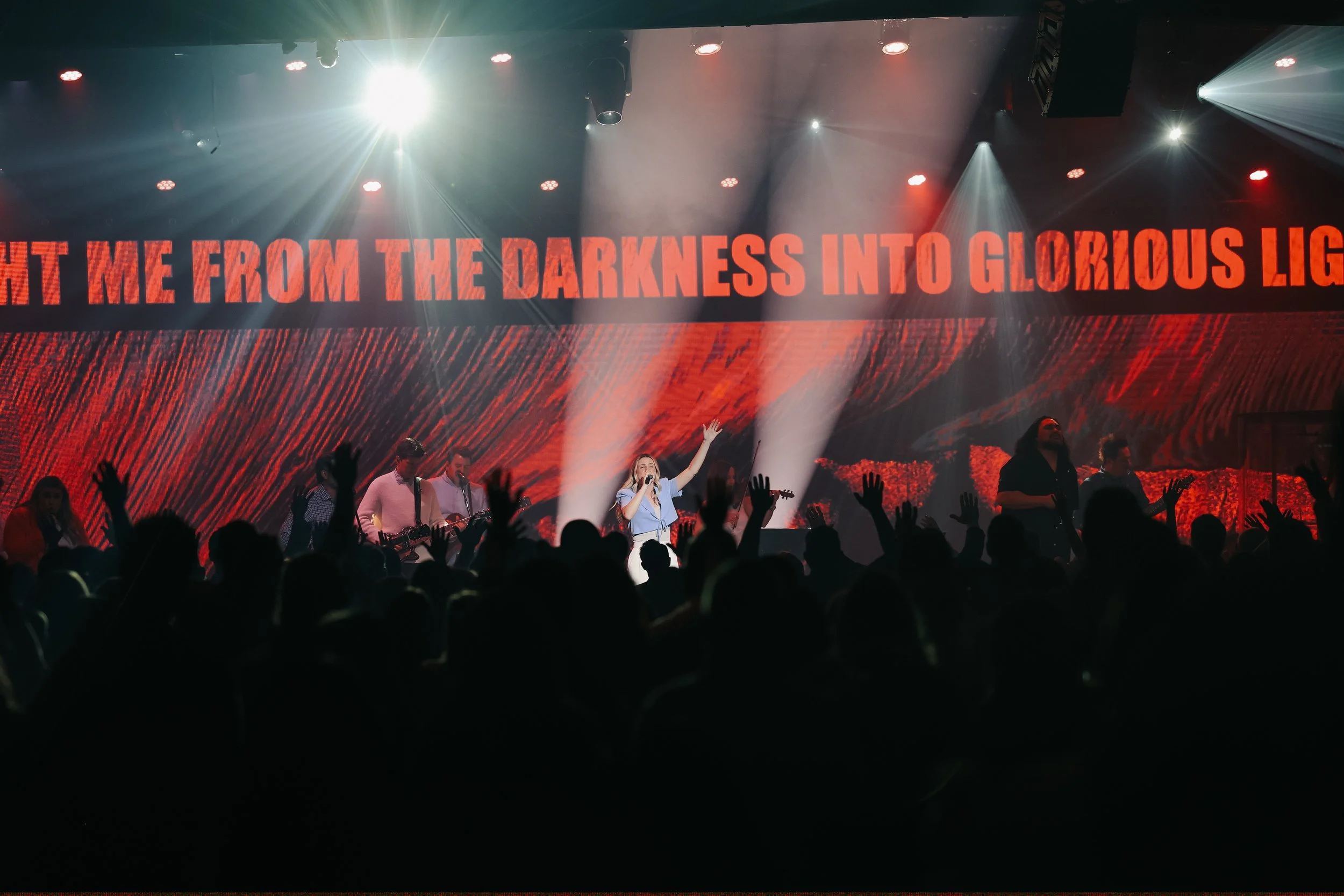 Concert scene with a singer and band performing on stage, audience raising hands, large red digital screen with the words 'Light me from the darkness into glorious light'