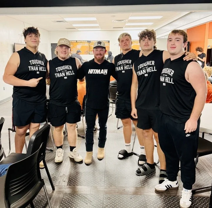Group of six young men posing together in a casual indoor setting, with some wearing black sleeveless shirts or t-shirts with athletic shorts. They are standing close, some with arms around each other, smiling or making relaxed gestures.