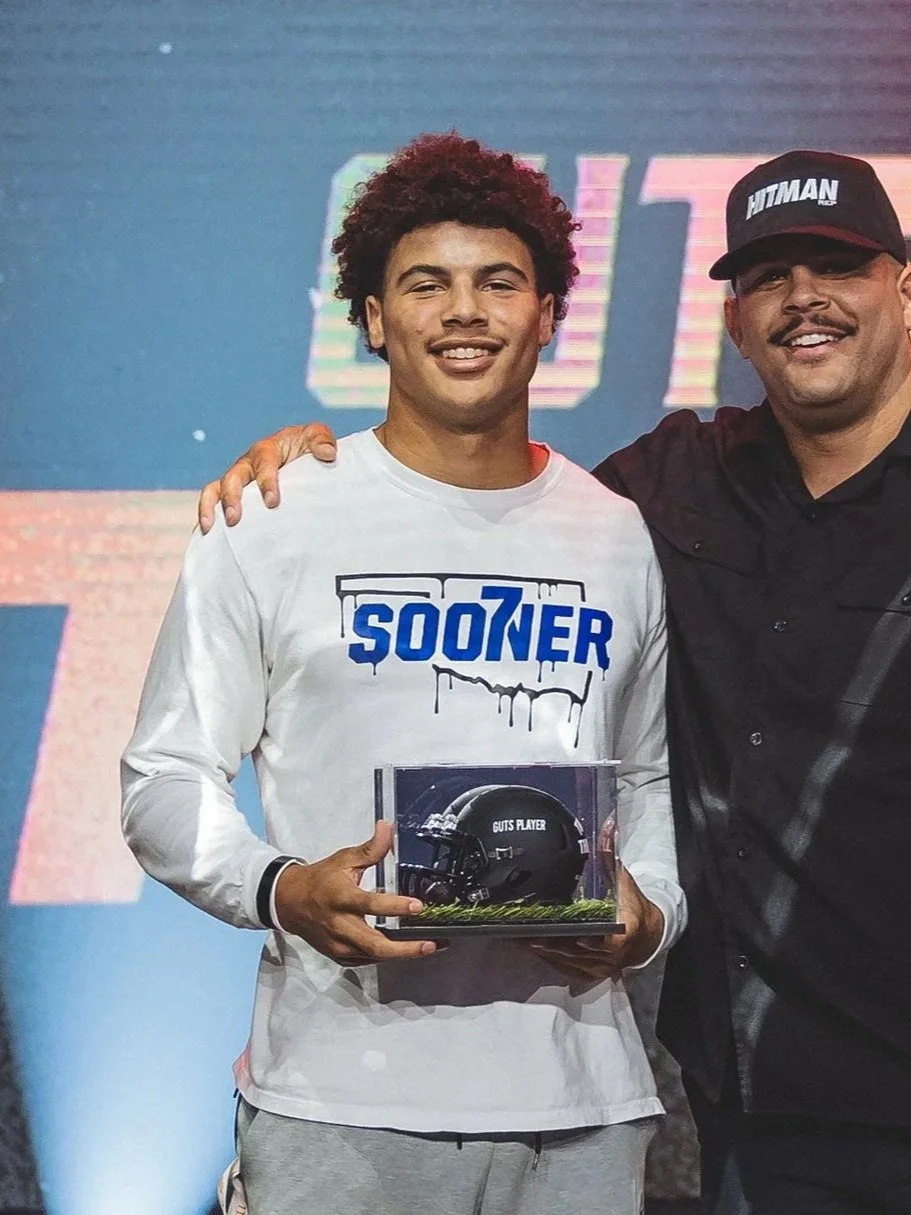 Young man with curly hair smiling while holding a plaque with a helmet labeled 'GUTS PLAYER,' standing next to an older man in a black shirt and cap, at an event with a digital screen behind them.