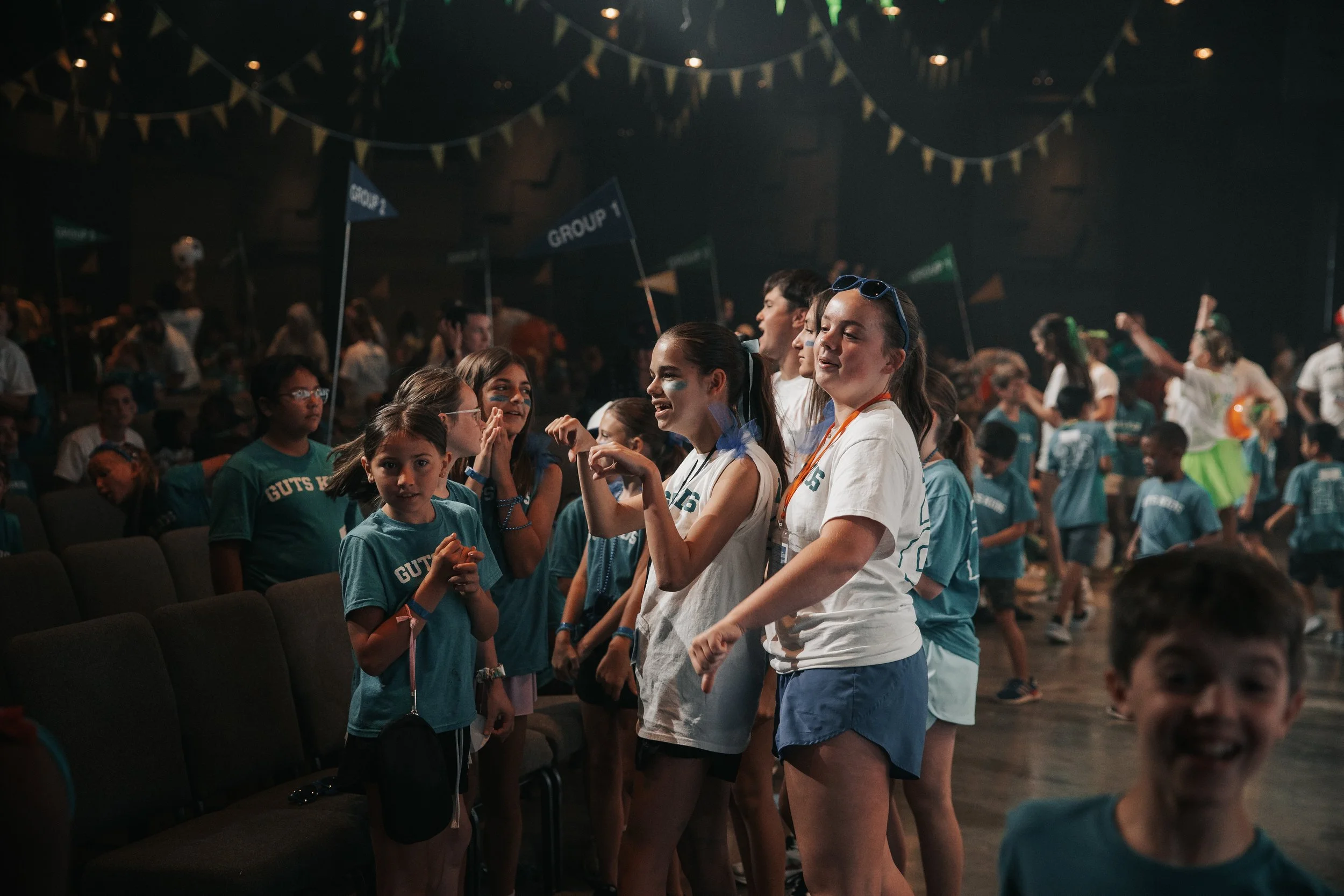 Group of children and teenagers standing in line inside a dimly lit auditorium or hall, some with face paint, wearing casual clothes including T-shirts with text, talking and interacting, while others are seated and watching.