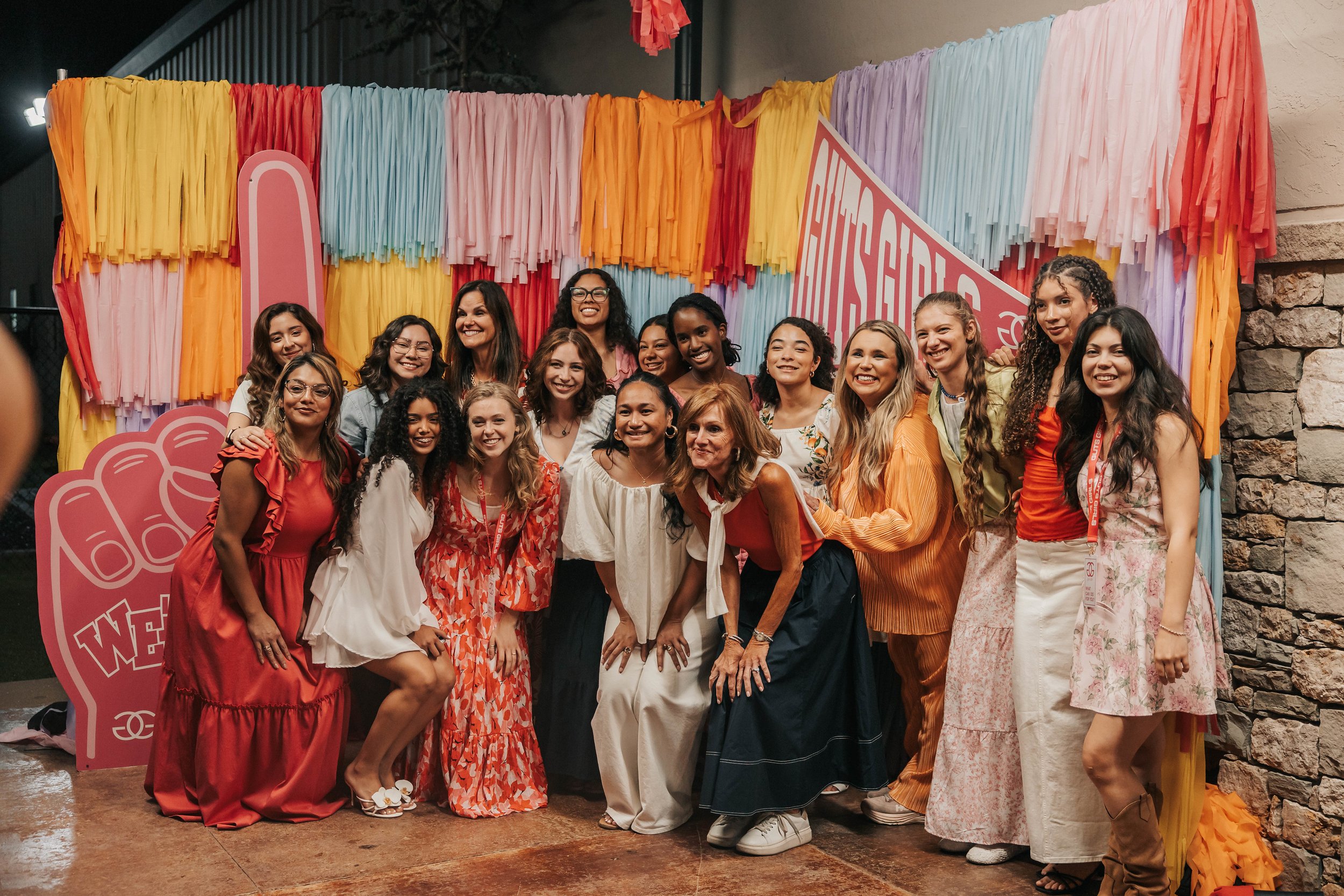 A group of diverse women and girls smiling and posing for a photo in front of colorful tassel backdrop with a hand sign graphic and promotional text for a women's event.