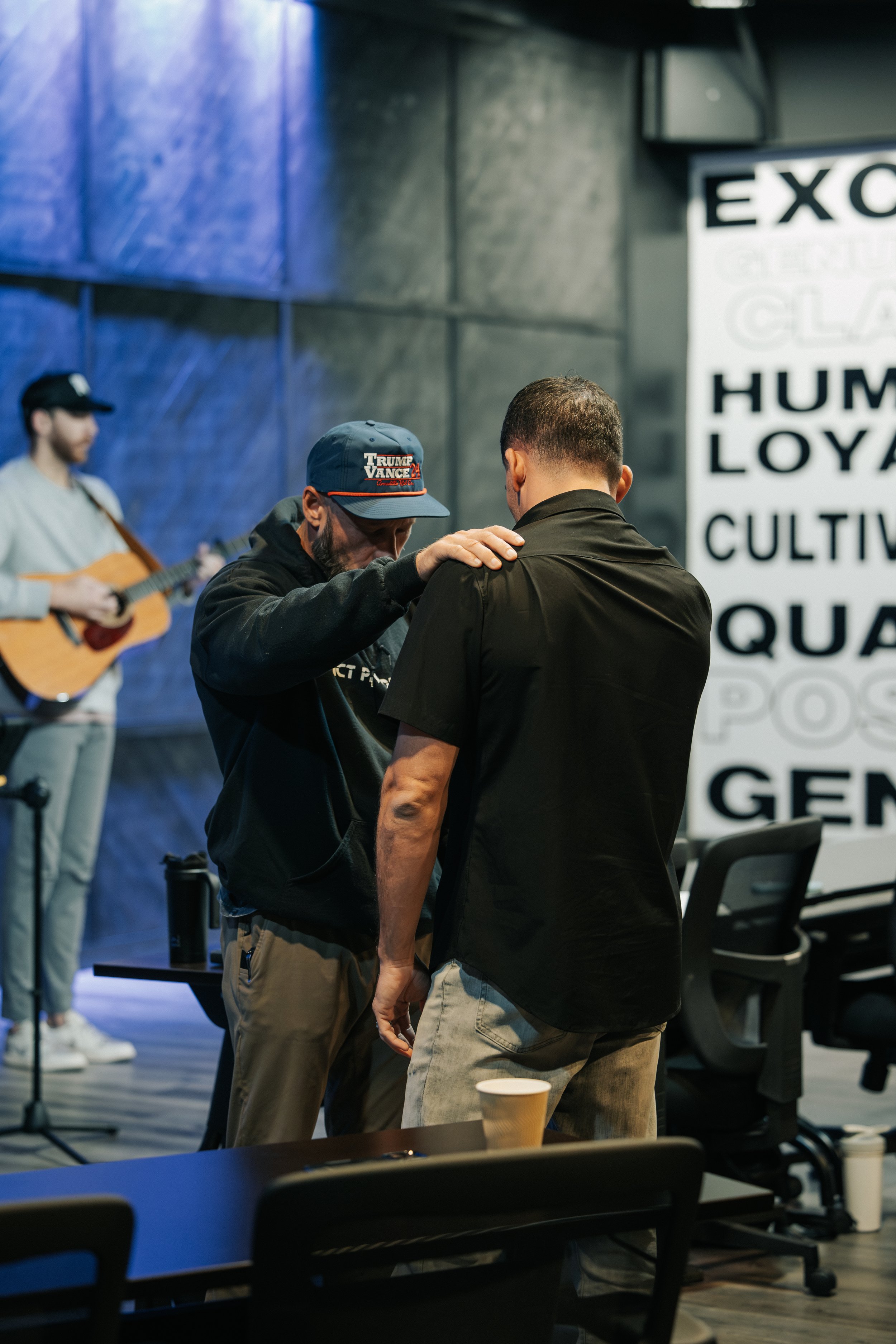 Two men are having a serious conversation in a room with a musician playing guitar in the background. The man on the left has a beard, wears a baseball cap and a hoodie, and has his hand on the shoulder of the other man, who has short hair and is dressed in a black shirt. There are chairs with cups on the table in the foreground and a large poster with words like 'EXO', 'LOYAL', and 'CULTIV' on the wall.