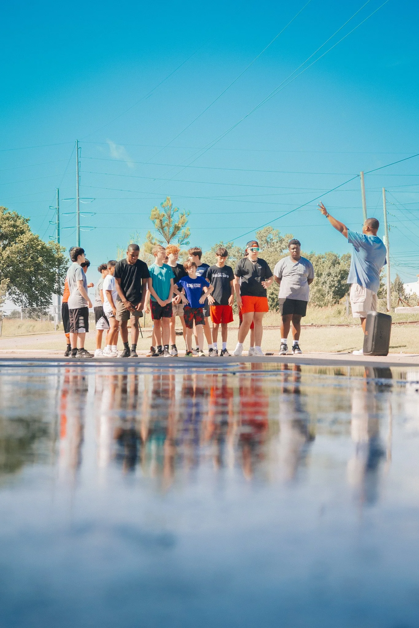 A group of children standing outdoors on a sunny day, listening to an adult who is speaking and gesturing with his right hand. The setting appears to be a park or sports field with trees, power lines, and a clear blue sky, with their reflection visible in a puddle in the foreground.