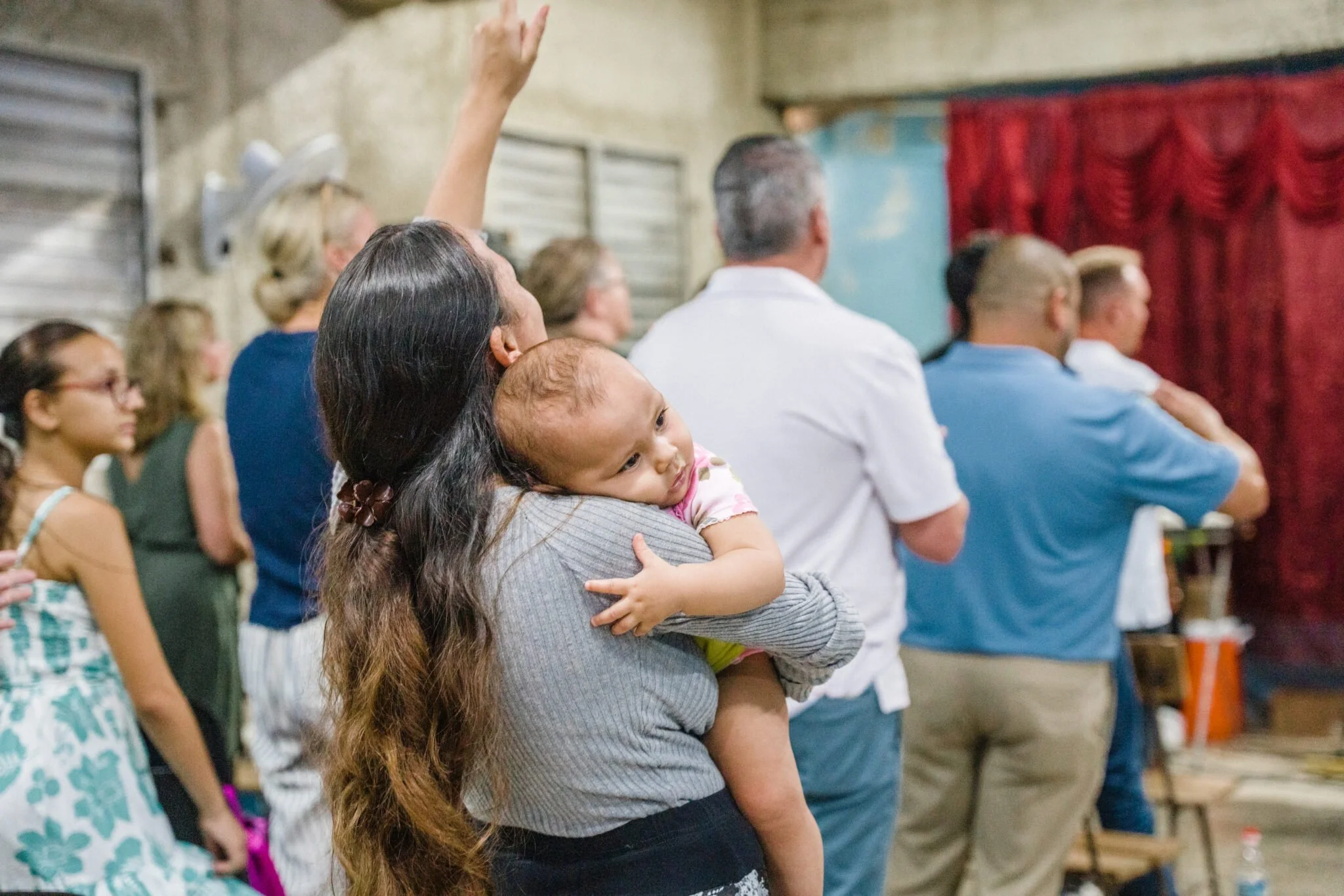 A woman holds a young child in a crowded room, watching a performance or event with red curtains in the background.