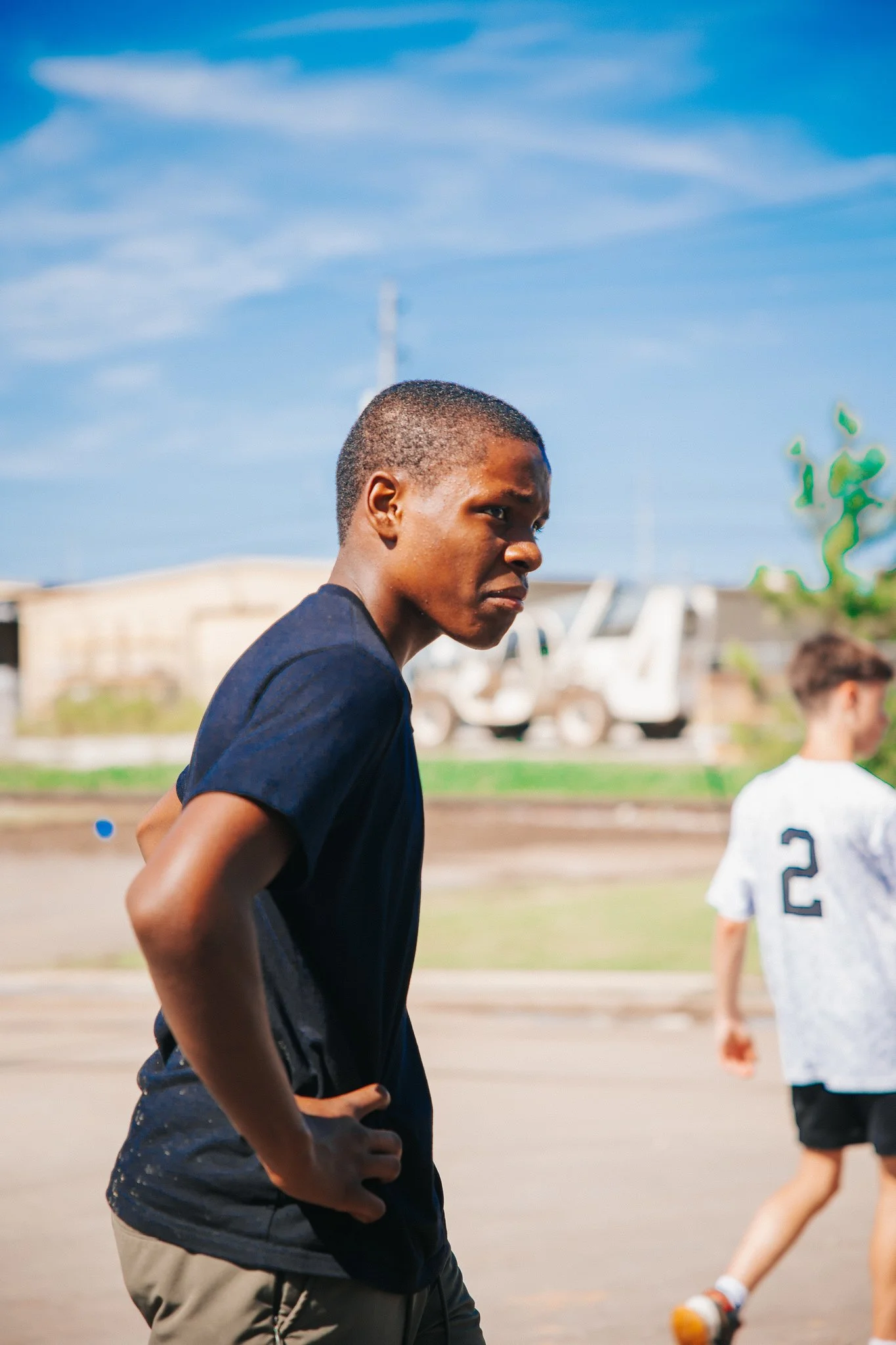 A young male athlete with a serious expression on his face, wearing a navy blue shirt, stands outdoors with his hands on his hips, during what appears to be a sports event or practice.