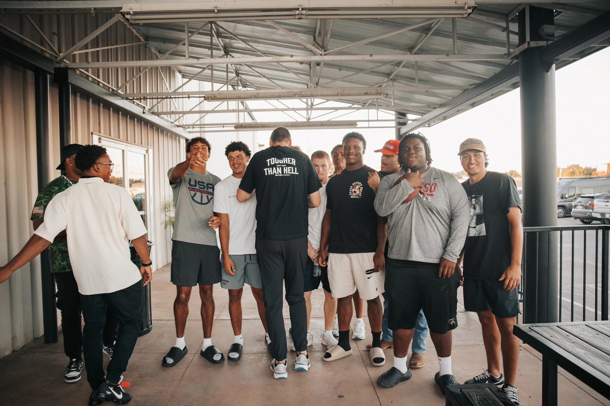 Group of young men standing together under a covered outdoor area, smiling and engaging with each other. They are casually dressed in T-shirts and shorts, some wearing caps, with a parking lot visible in the background.