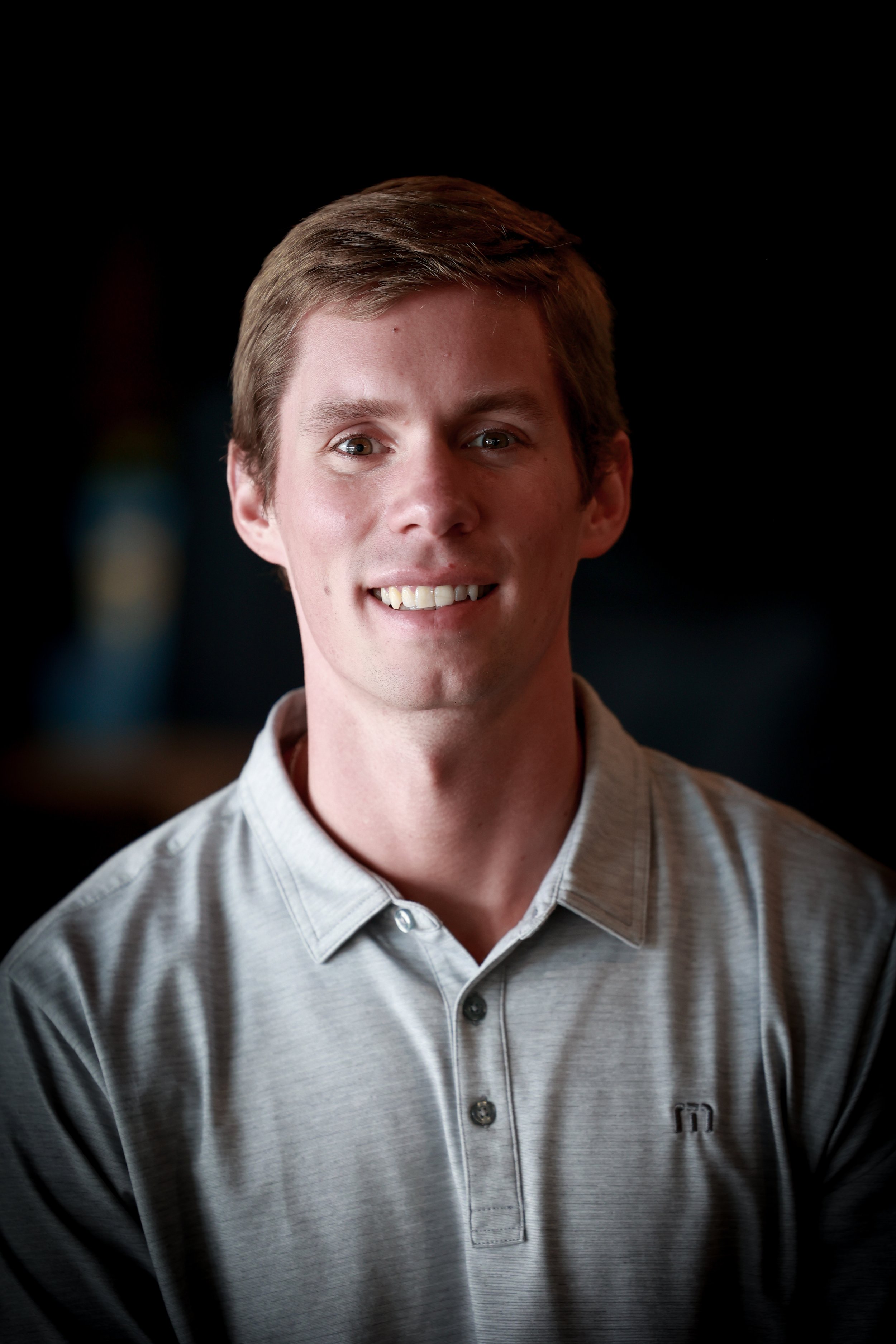 Portrait of a young man with light brown hair smiling, wearing a light gray collared shirt, against a dark background.