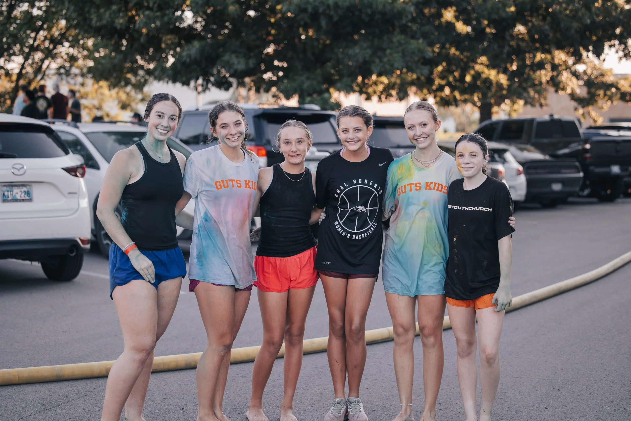 Six young girls standing arm in arm outdoors after a sports event, with cars and trees in the background.