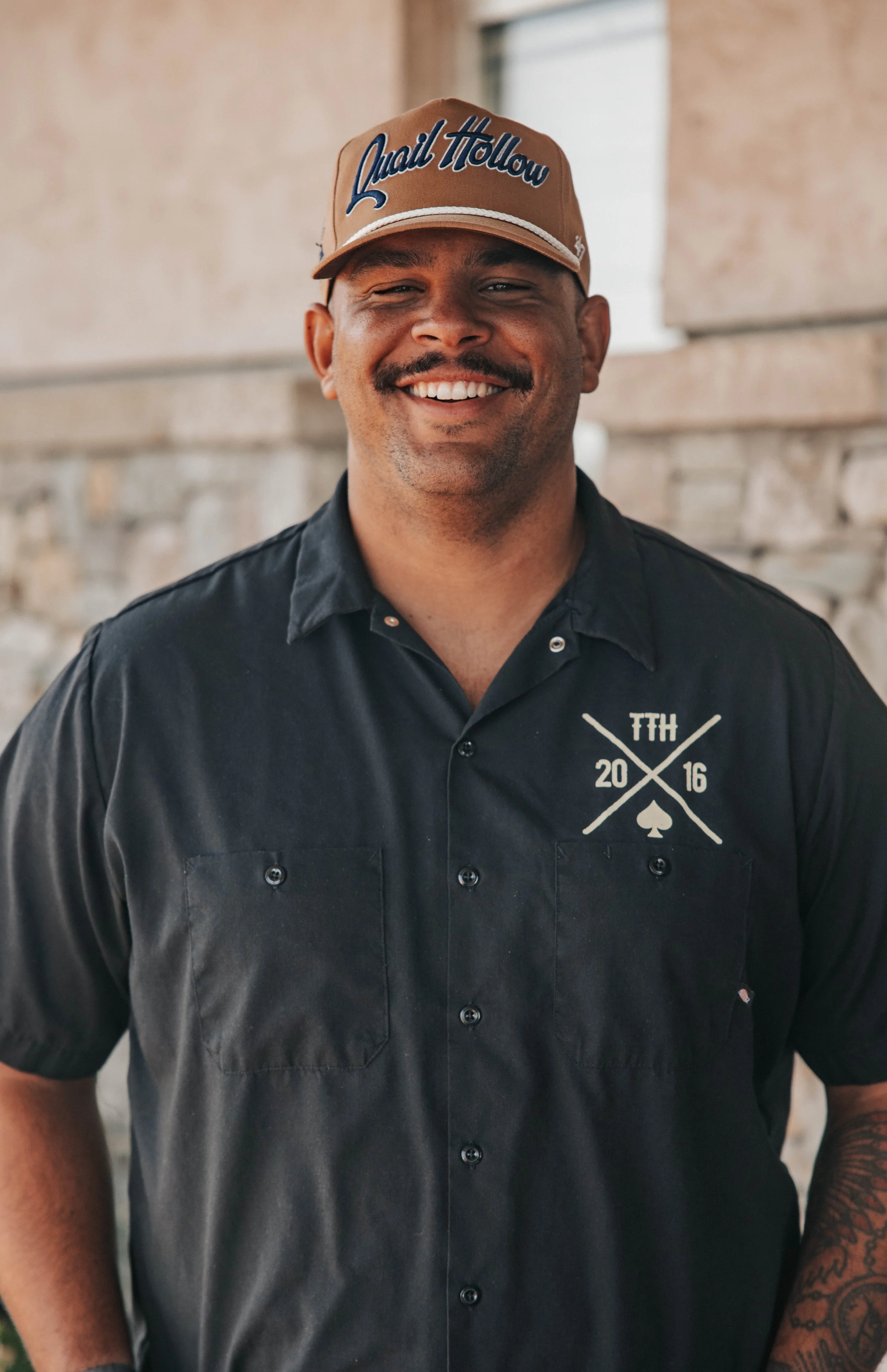A smiling man wearing a tan cap with 'Loyal Hollow' written on it, a black short-sleeve button-up shirt with a crest design, and standing outdoors in front of a building.