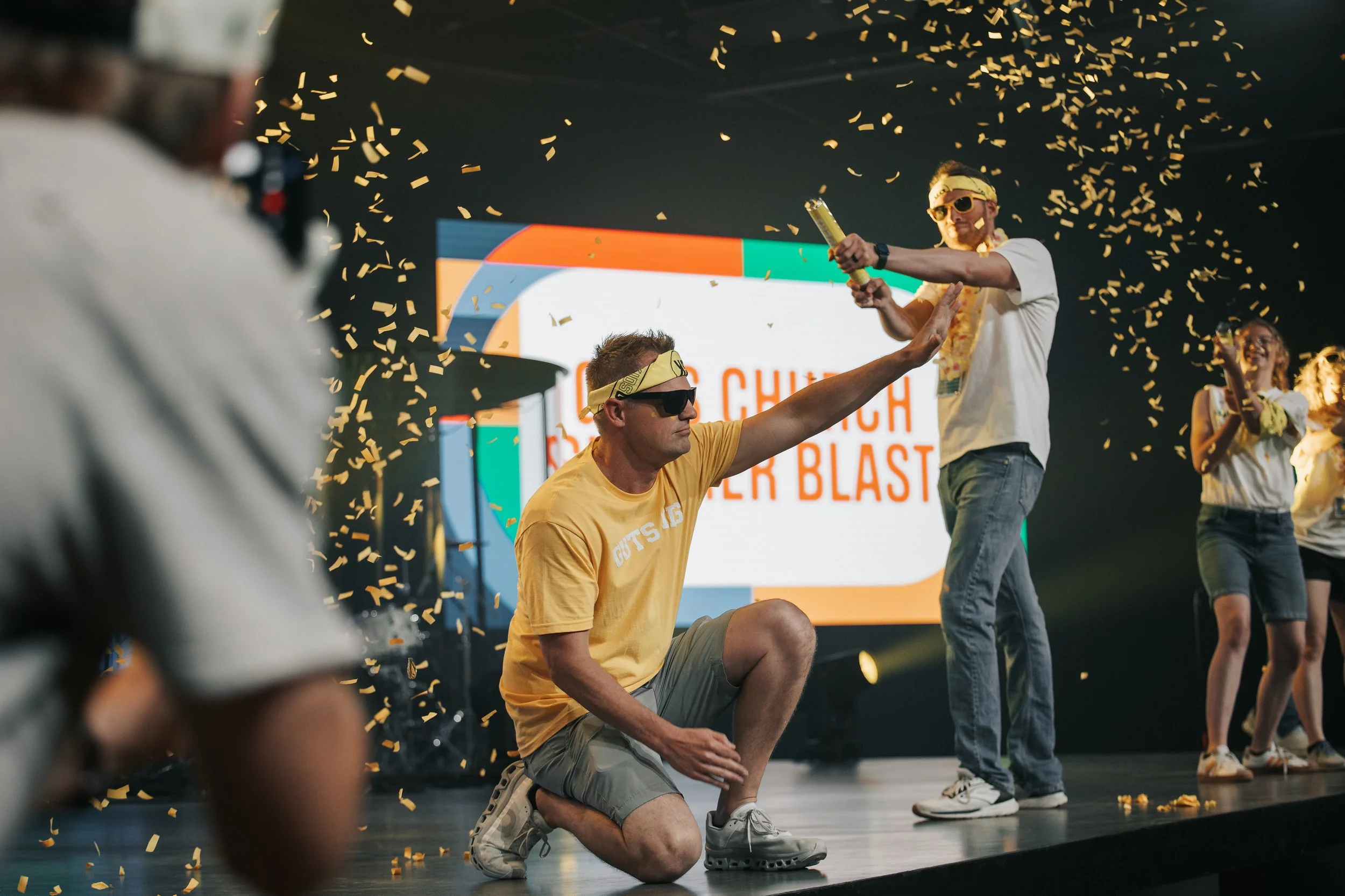 People celebrating on stage with confetti, one kneeling and three standing, in front of a large screen with colorful graphics.