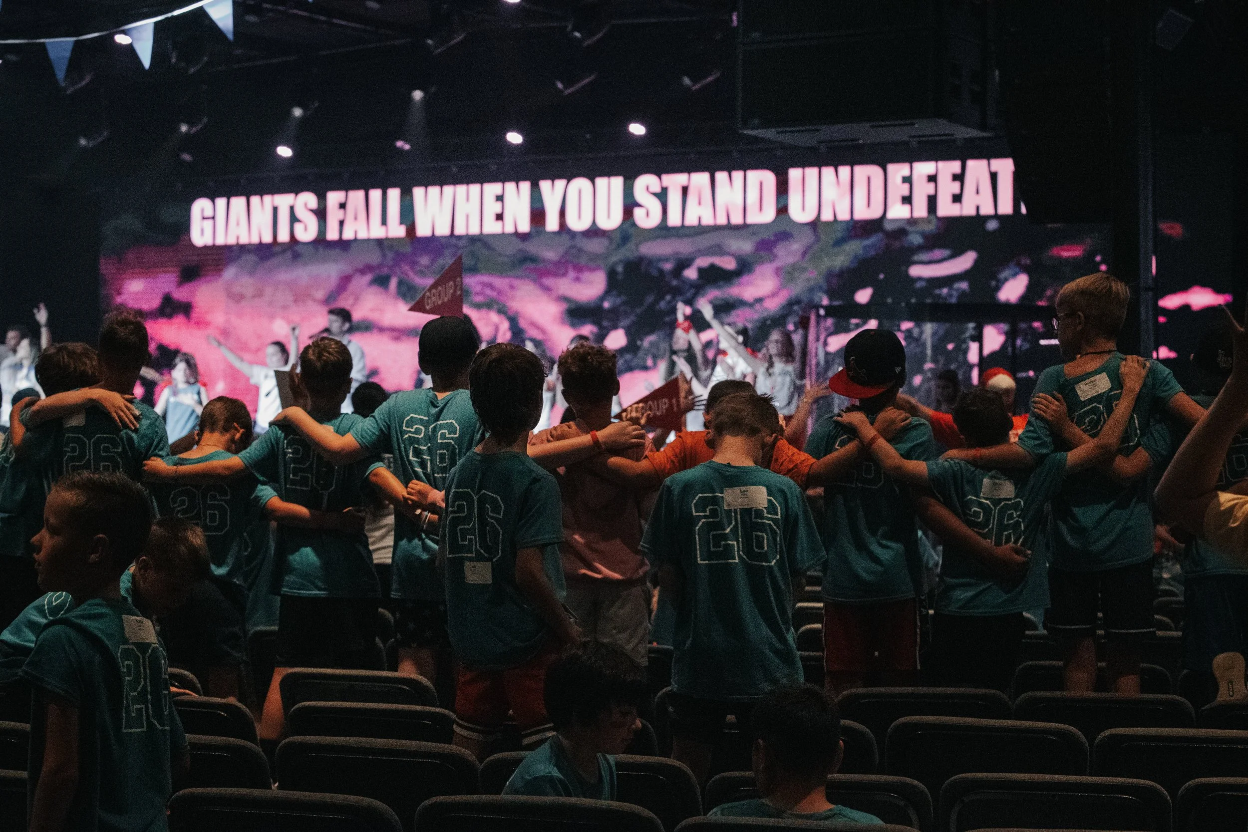 A group of children wearing teal shirts, standing in a circle with arms around each other's shoulders, facing a stage where a screen displays the message 'Giants fall when you stand underfeat'. The stage and audience are in a dark room, with some chi