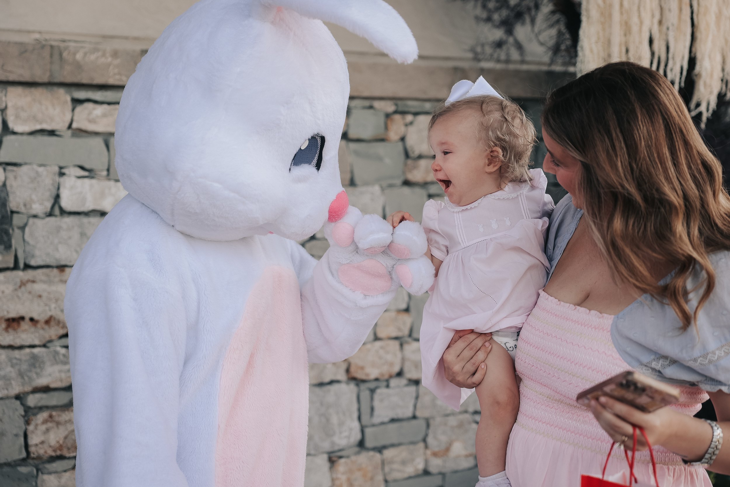A woman holding a smiling young girl in her arms and interacting with a person in a white bunny costume against a brick wall background.