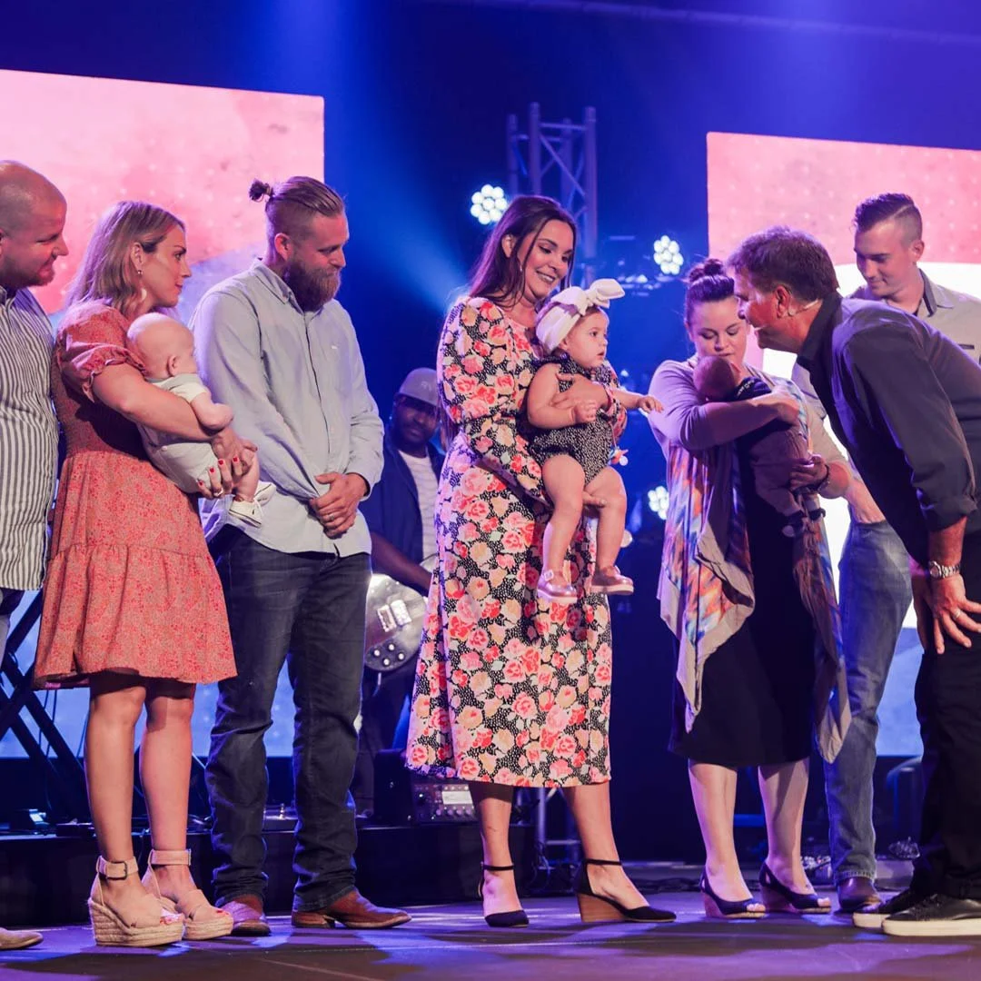A group of adults and children on stage during a ceremony, with some holding babies and others smiling and interacting.