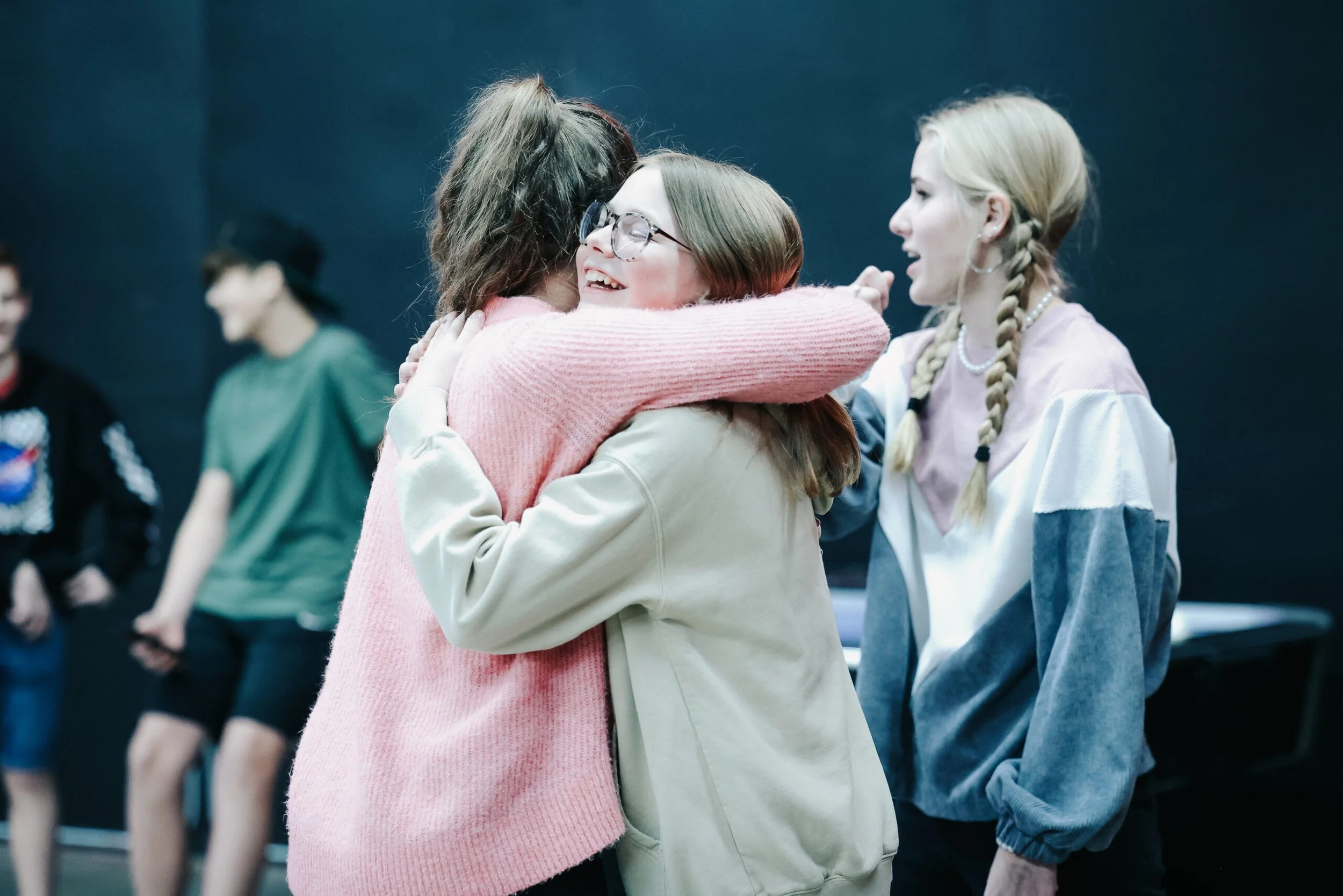 Three young women hugging and smiling at each other in a casual setting, with two other people in the background.
