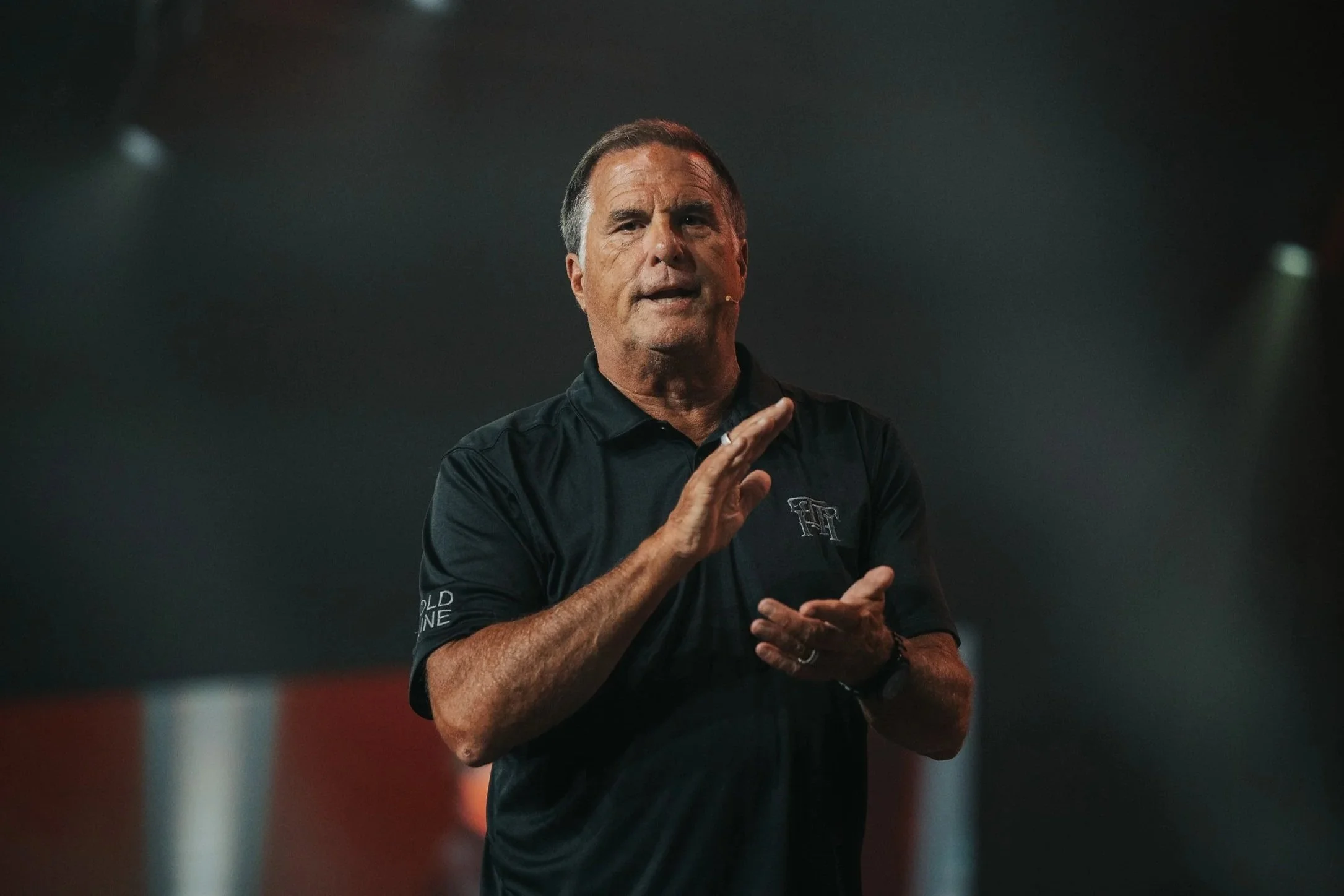 A man wearing a black shirt and jeans standing on stage giving a presentation to an audience in a dark setting.
