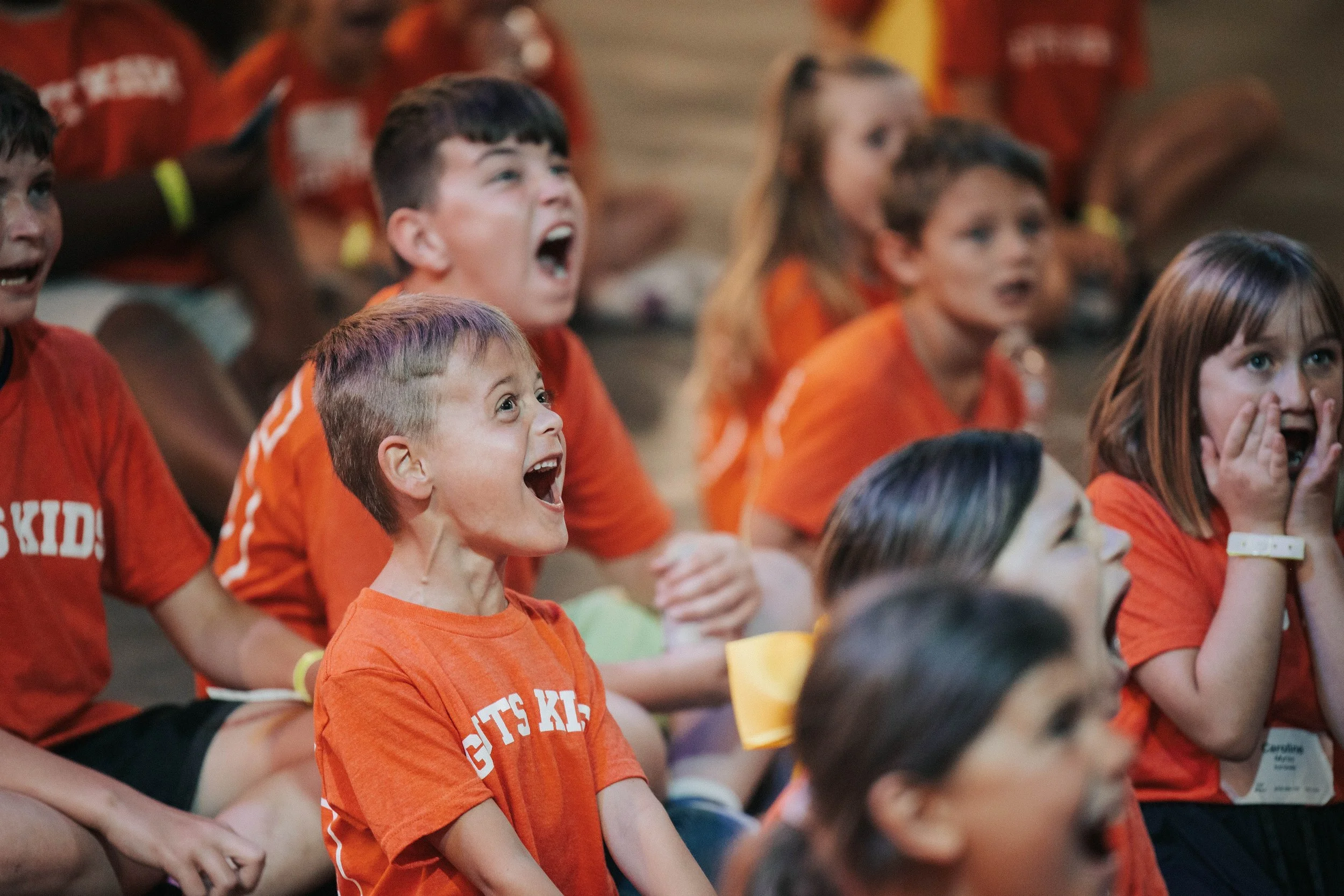 Group of children wearing orange shirts, sitting and reacting with excitement and surprise.