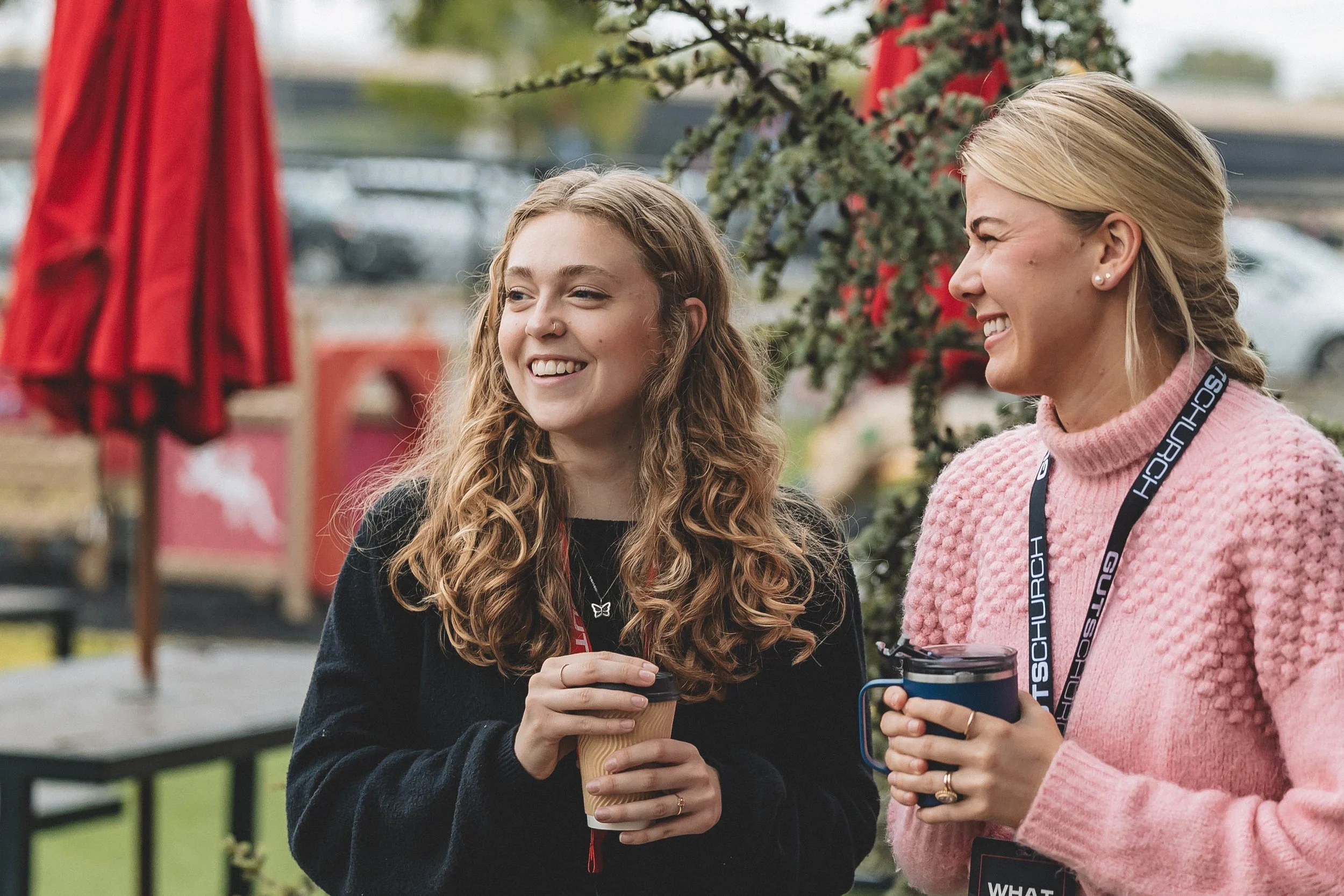 Two women smiling and talking outdoors, holding coffee mugs, with red umbrellas and greenery in the background.