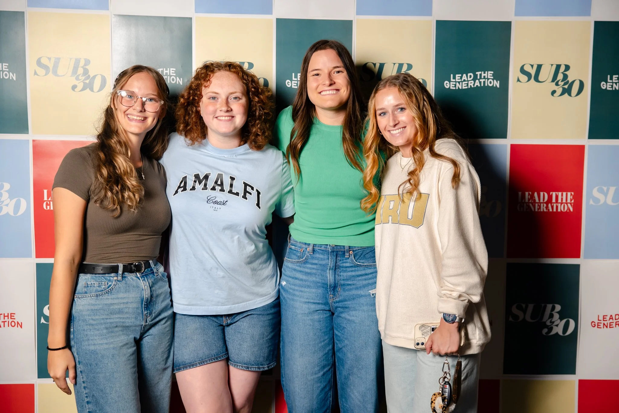 Four young women standing together in front of a colorful branded backdrop, smiling.