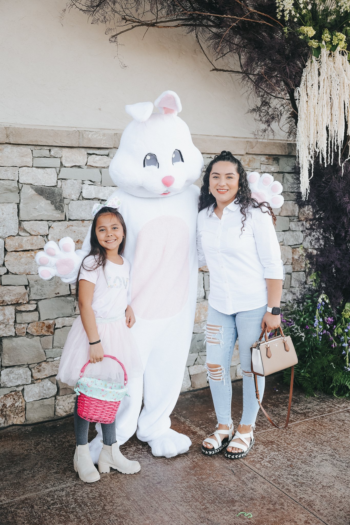 A young girl and a woman standing next to a person dressed in a white bunny costume with big, expressive eyes, a pink nose, and large paws, against a stone wall and greenery background.