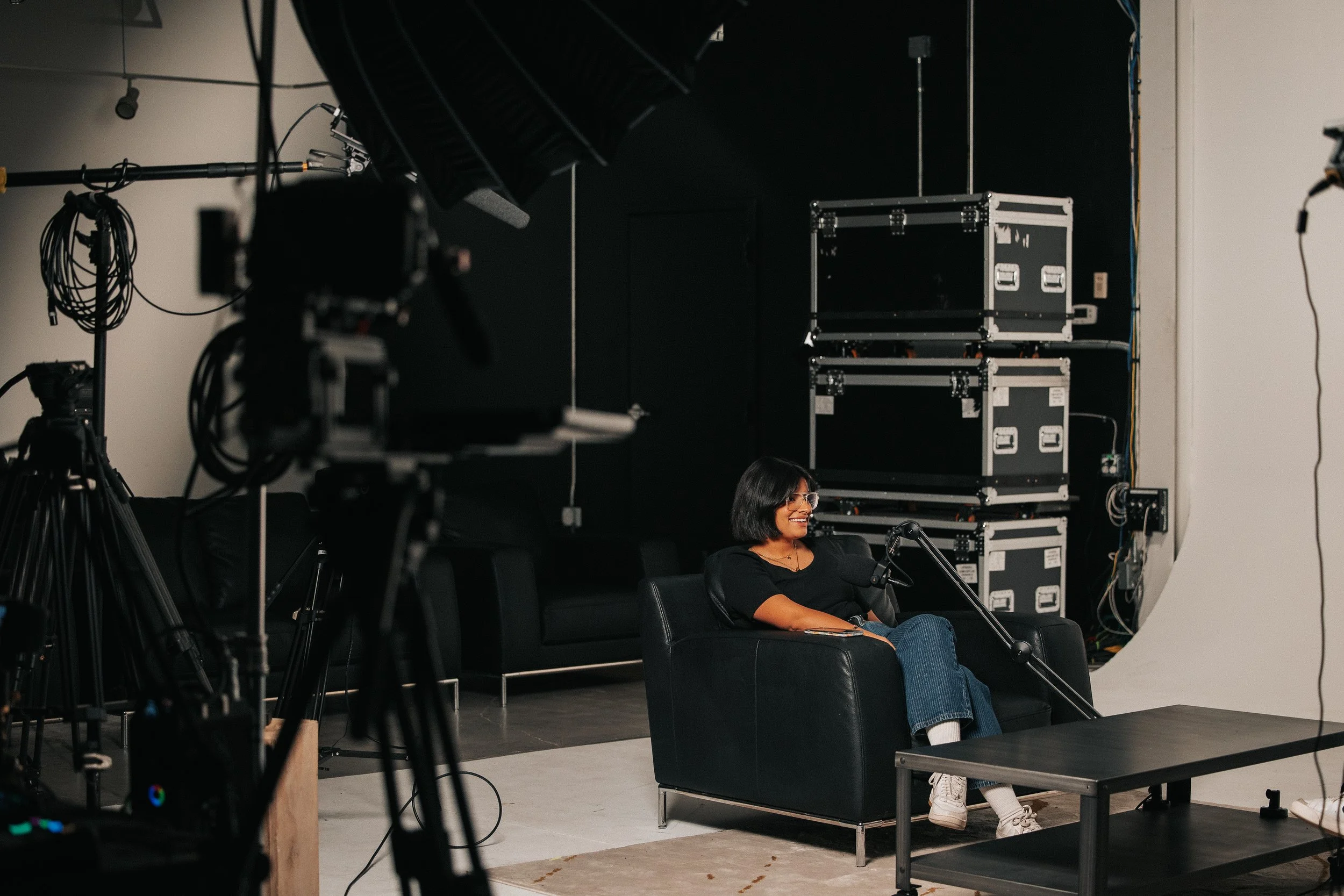 A woman sitting on a black couch in a studio or recording setting, smiling, with professional equipment, cameras, lighting, and audio gear around her.