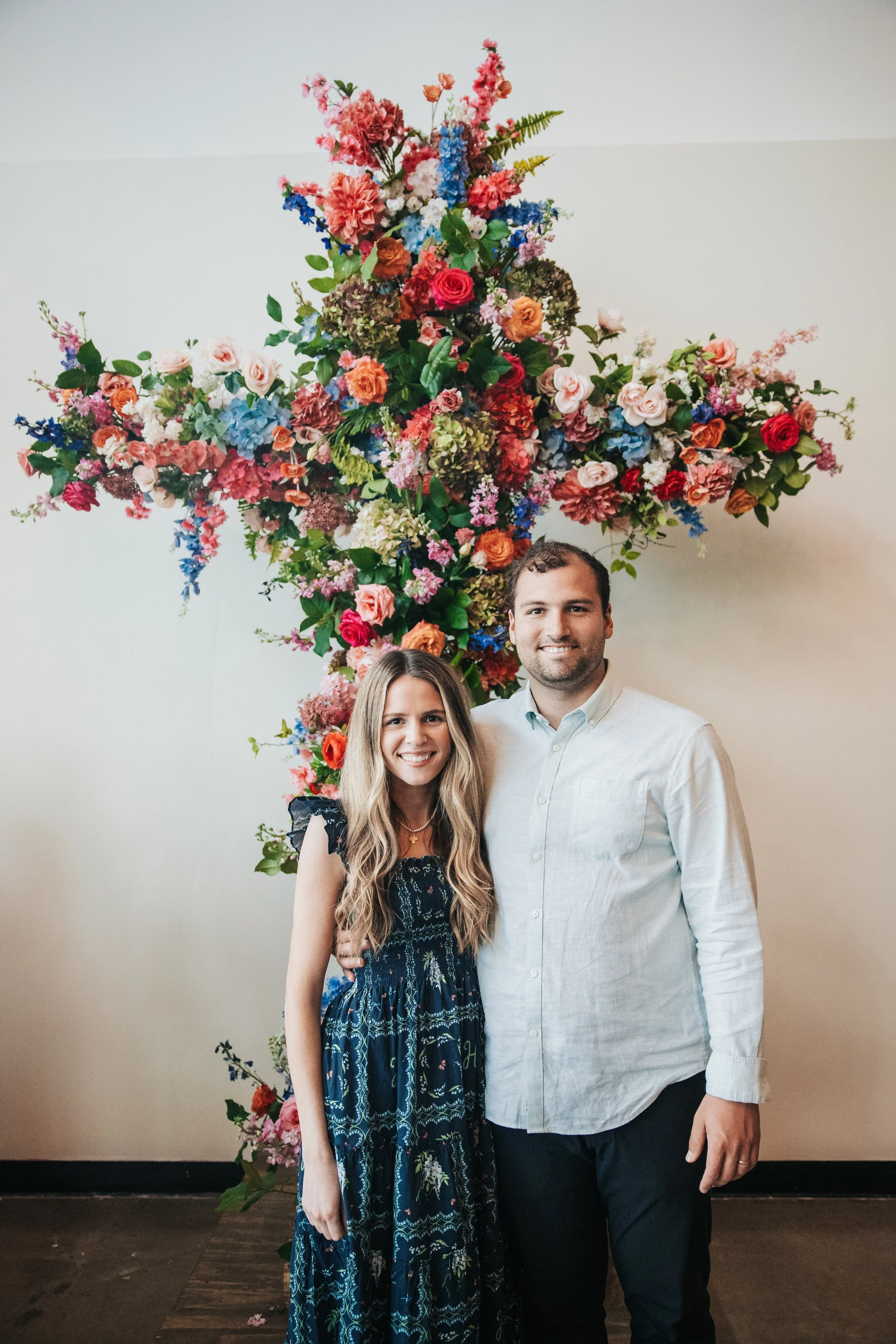 A smiling woman and man standing in front of a large colorful floral cross.