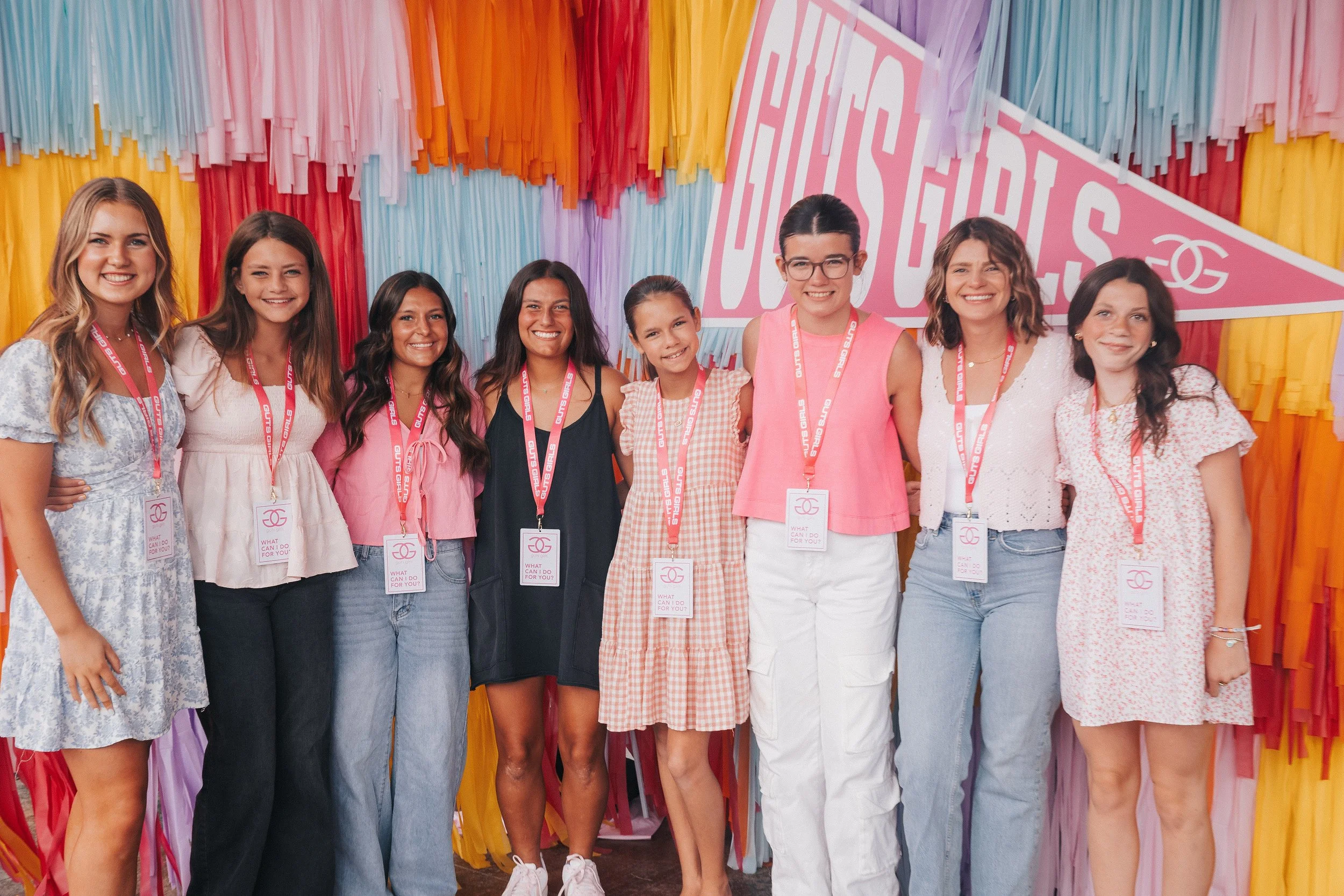 A group of nine young women standing together in front of a colorful backdrop with pink, purple, yellow, and orange ribbons, smiling and posing for a photo at an outdoor event.