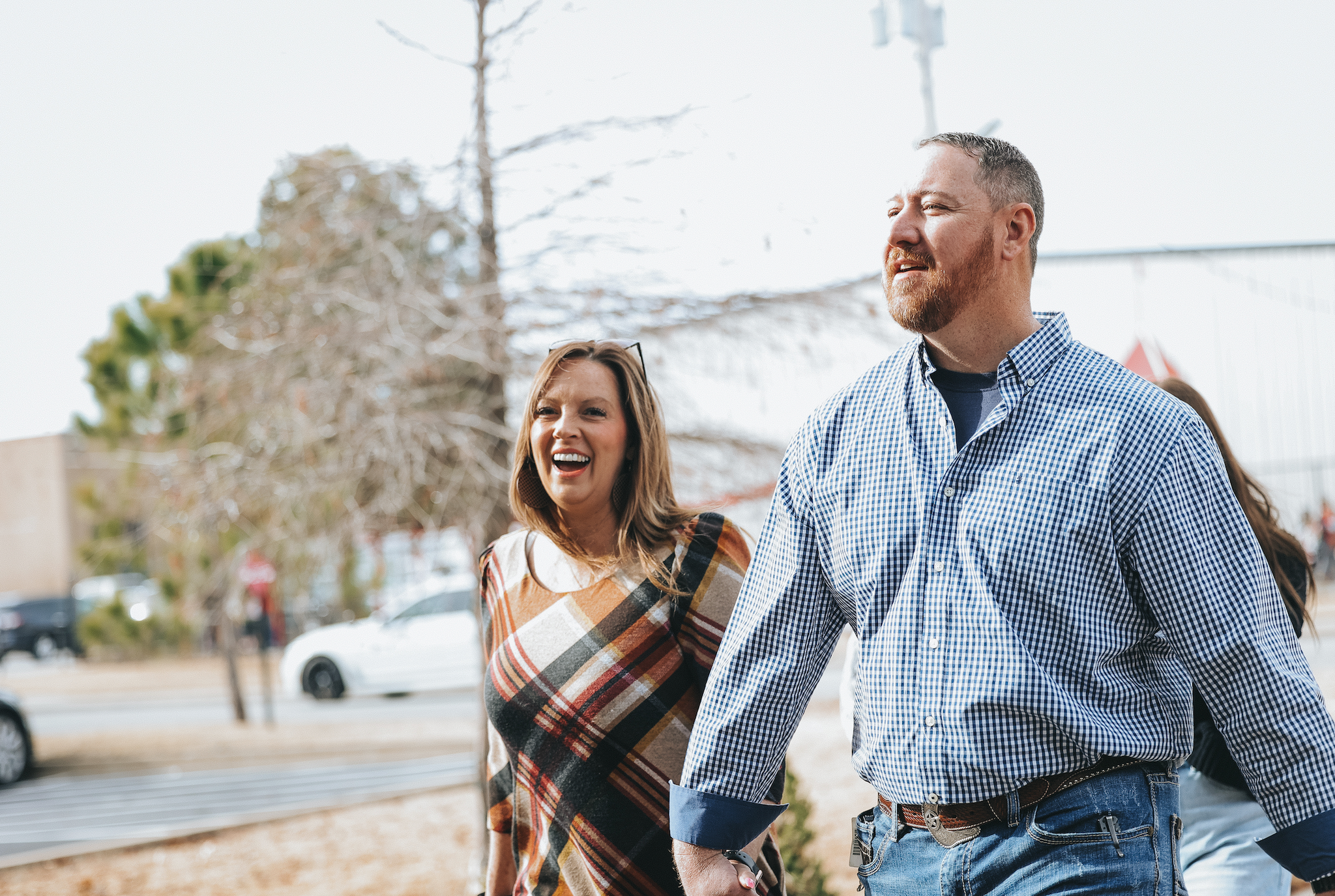 A group of smiling people walking outdoors on a cloudy day, with a woman in a plaid shirt and a man in a checkered shirt in the foreground.